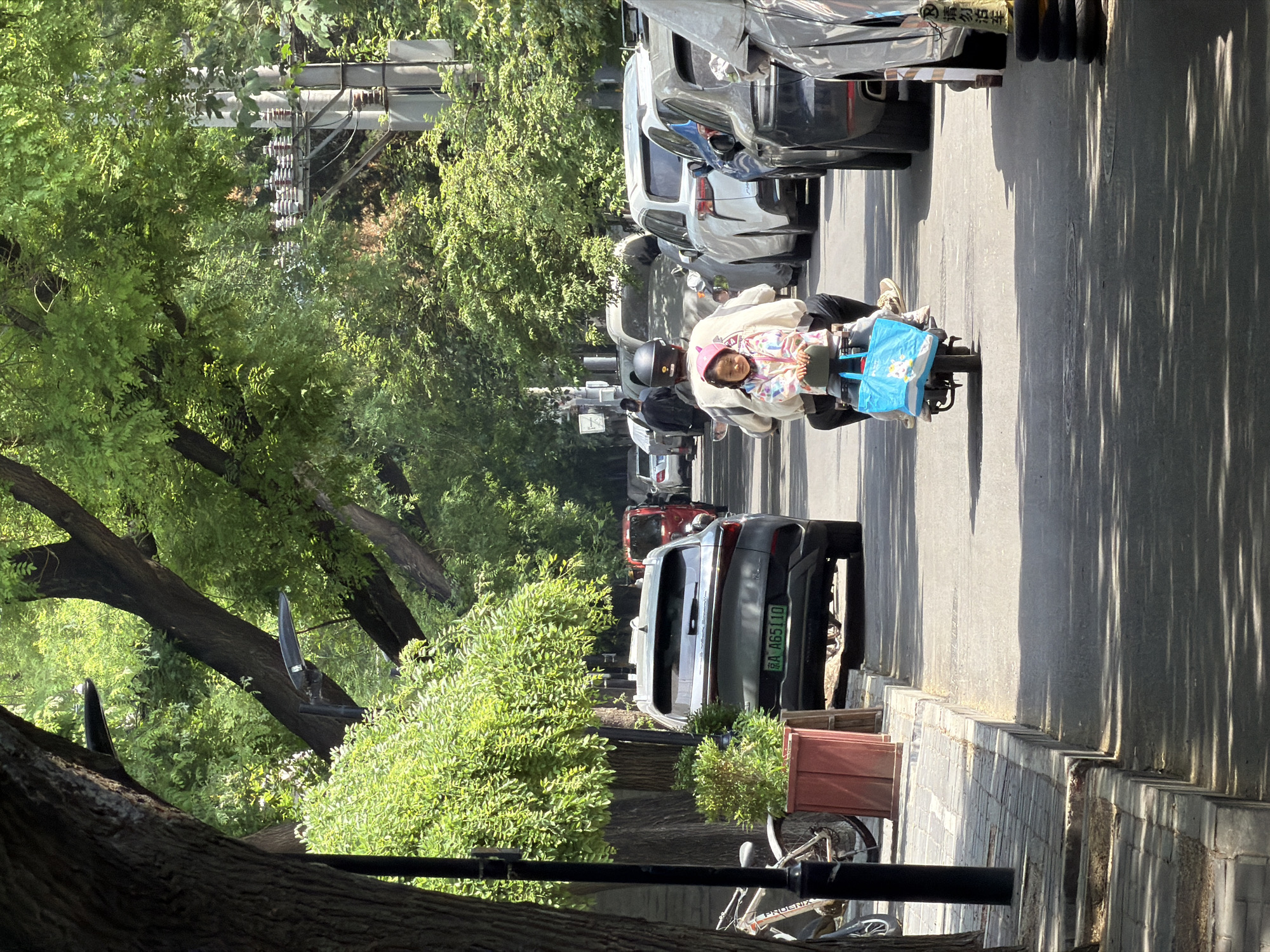 A street scene shows a person with a child on a small motorcycle. The person is wearing a dark helmet, and the child is wearing a pink helmet. A blue bag is on the front of the motorcycle. The street is lined with parked cars. The license plate number of a car is 京A A65110. Trees line the street, casting shadows on the sidewalk. In the distance, power lines are visible. The scene appears to be in the Dongcheng District of China.