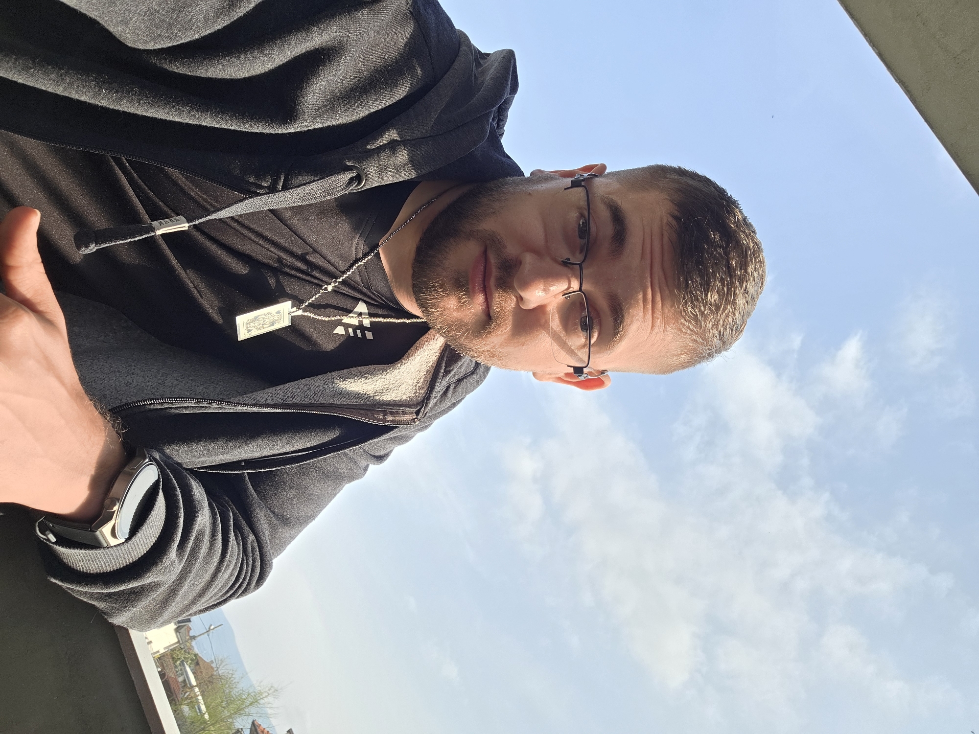 A man wearing glasses, a black t-shirt, and a dark grey hooded jacket stands on a balcony with a smartwatch on his left wrist. He is looking down at the camera and giving a thumbs-up. A silver chain necklace with a rectangular pendant is visible. The background features a bright blue sky with scattered clouds. In the distance, residential buildings and trees can be seen. The location is identified as Yagodovo, Bulgaria.