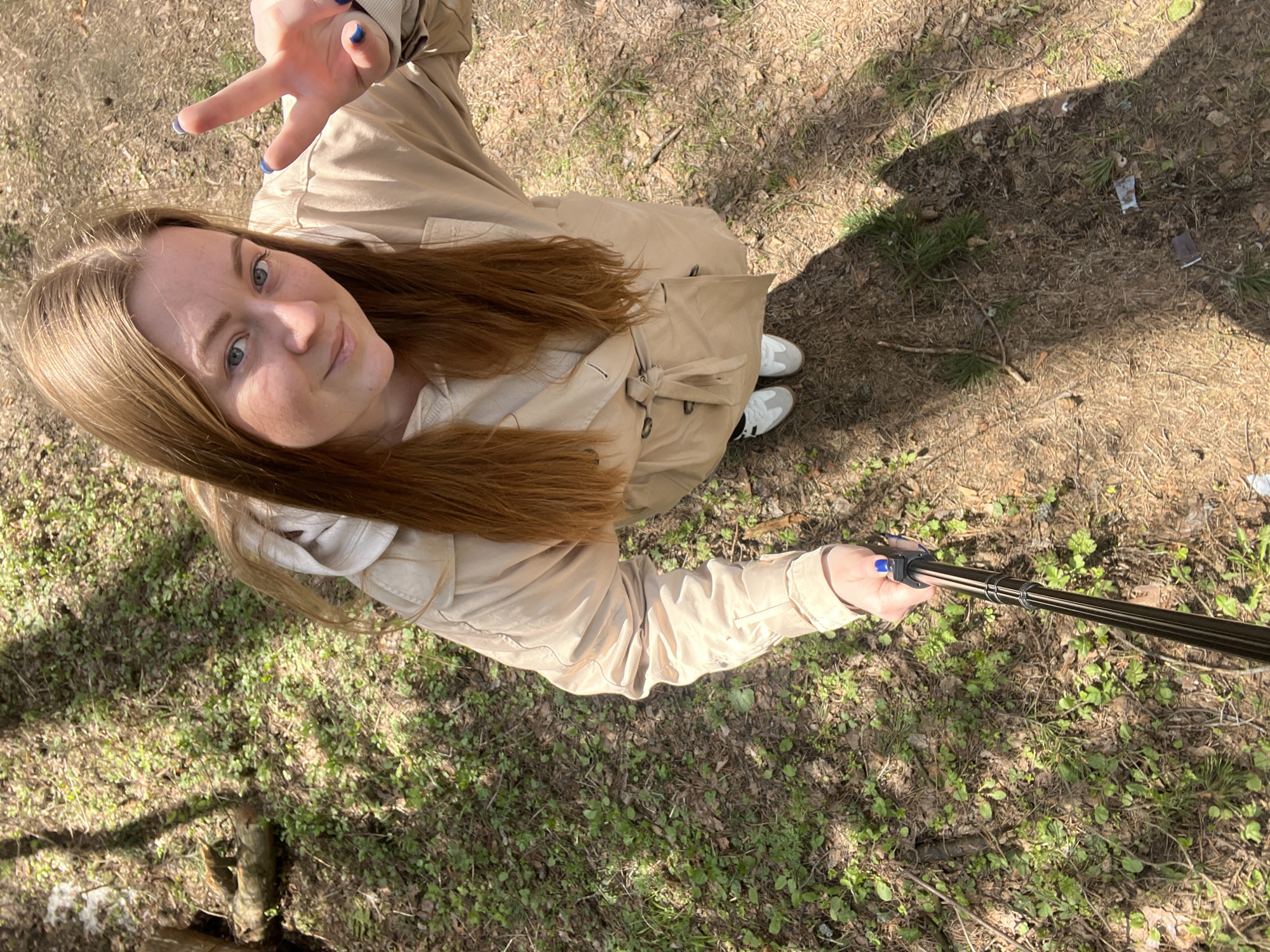 A woman with long auburn hair is taking a selfie in what appears to be a park or wooded area. She is wearing a beige trench coat and white sneakers. She is holding a selfie stick in her right hand and making a peace sign with her left hand. The ground is covered in dirt, grass, and fallen leaves. The location is Раздолье, Russia.