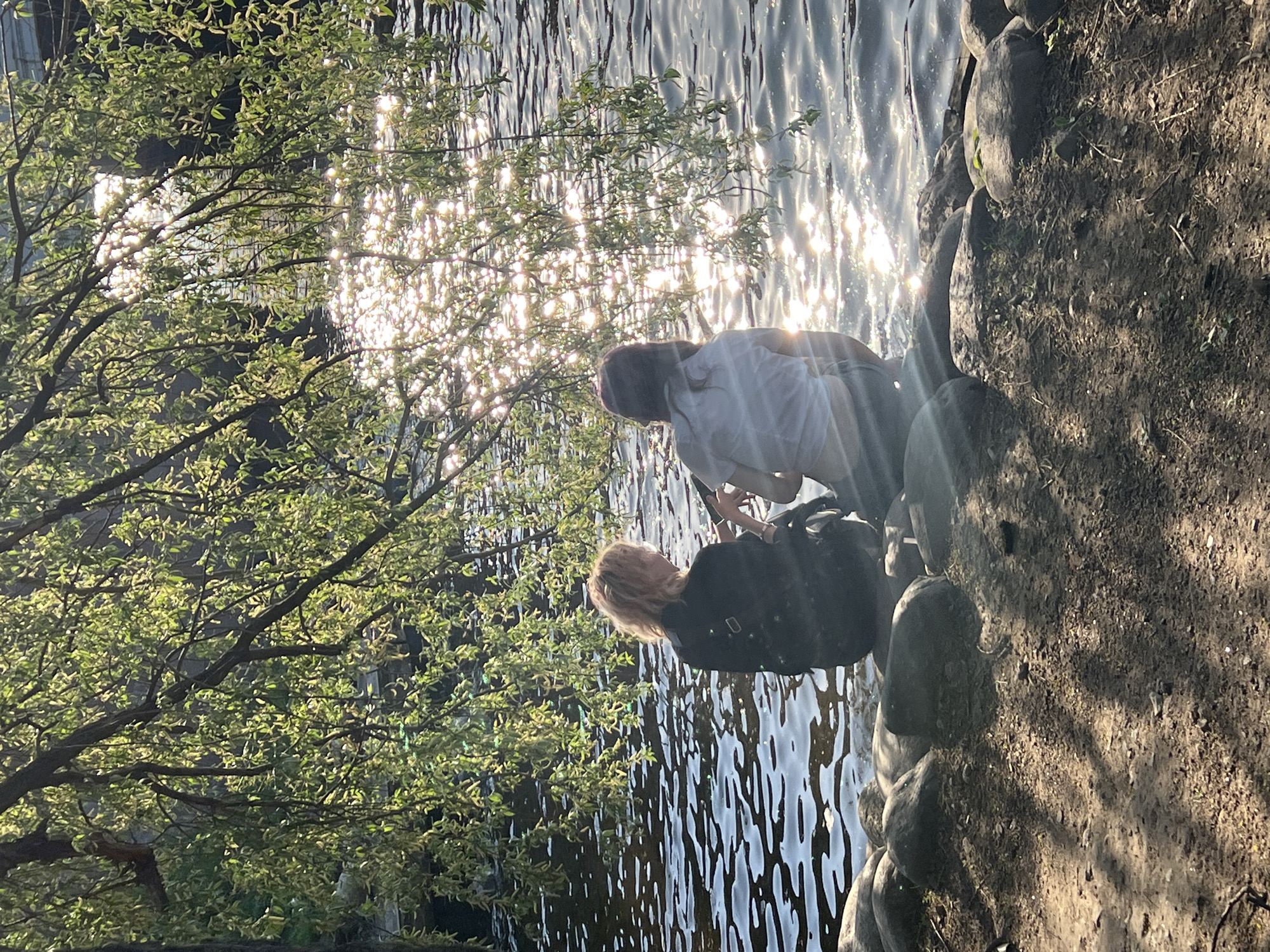 The image depicts two individuals near a body of water, possibly a canal or river, in what appears to be a park setting. The location is likely Saint Petersburg, Russia, given the architectural style and general landscape.
The first person, facing away from the camera, is wearing a dark jacket and possibly carrying a dark-colored bag on their shoulder. The second person, closer to the water's edge, is wearing a white top and light-colored pants. They appear to be interacting with something in or near the water.
The surrounding environment includes foliage, with a tree on the left side of the image providing a partial canopy. The water surface reflects sunlight, creating bright highlights. The edge of the water is lined with rocks and soil. The ground is primarily composed of earth and is partly in shadow.