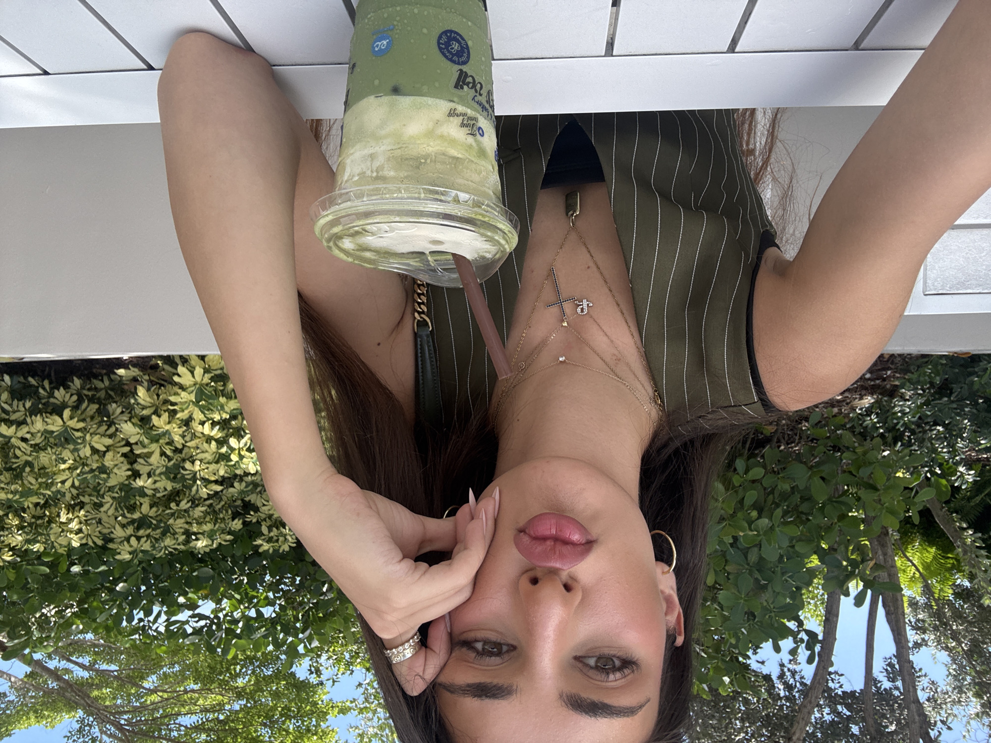 In Hallandale Beach, Florida, USA, a young woman with long, dark hair and makeup sits at a white table outdoors. She wears a green, pinstriped vest over a black top, adorned with several gold necklaces, including one with a cross pendant and another with a rectangular tag. A gold hoop earring dangles from her left ear, and she sports multiple rings on her right hand, one with a checkered pattern. Her right arm rests on her cheek, and her lips are puckered. In front of her is a clear plastic cup filled with a green beverage, likely a matcha latte, with a brown straw. The background features lush green foliage and trees under a bright blue sky.