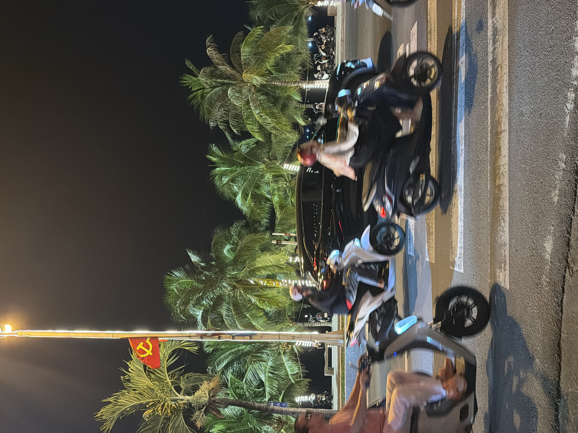 The image depicts a nighttime scene on a street in Nha Trang, Vietnam. A red flag with a yellow hammer and sickle, likely the flag of Vietnam, flies from a tall pole adorned with lights. Palm trees with illuminated trunks line the street. Several individuals are riding scooters on a road marked with a pedestrian crosswalk. One scooter in the foreground is a light color, and another in the middle ground is dark. A black car is visible behind the scooters. The overall atmosphere is that of a busy urban environment at night, with a tropical ambiance due to the presence of palm trees.