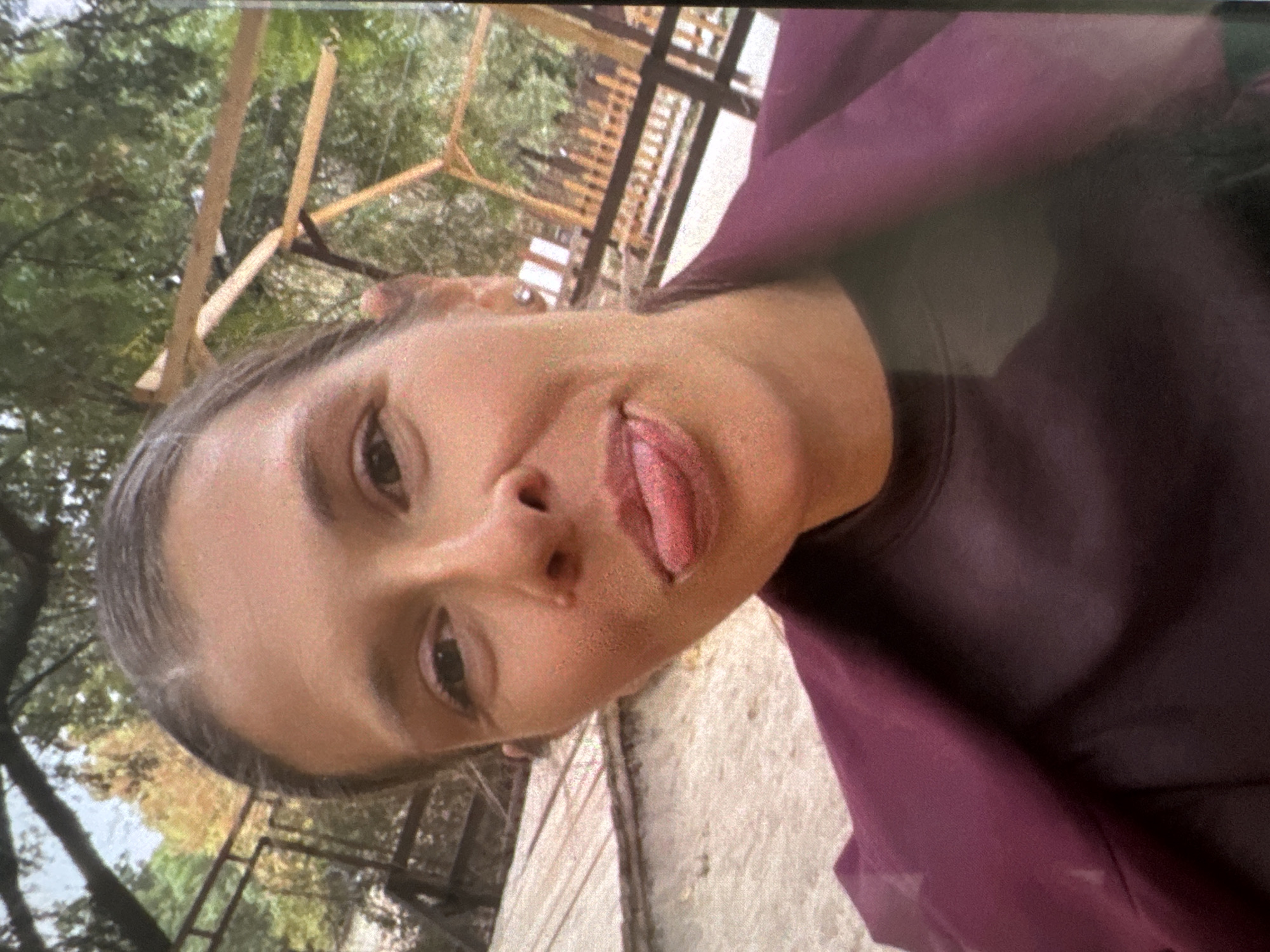 An adult female, approximately 30-40 years old, is depicted in a close-up outdoor photograph taken in Vratsa, Bulgaria. Her dark brown hair is pulled back, revealing a visible central part and some lighter hair strands near the forehead. Her skin tone is fair to medium, with a small mole visible superior to the right corner of her upper lip and another small mole on the right side of her nasal bridge. Her dark brown eyes are open and directed towards the camera. Her lips are pursed, with her tongue partially extended from her mouth, indicating a playful expression. She is wearing a dark, possibly deep maroon or plum-colored long-sleeved top beneath an open blazer or jacket of a slightly lighter purple or plum hue. Small stud earrings are visible in both ears. The background features light-colored sandy or dirt ground to the subject's left and dense green foliage from trees, interspersed with patches of yellow and orange autumnal leaves. Visible wooden structures include a dark railing or fence on the left side and a light-colored wooden pergola or open-frame structure with horizontal and diagonal beams on the right side. Additional wooden fencing and structures are discernible in the deeper background, consistent with an outdoor recreational or park environment.