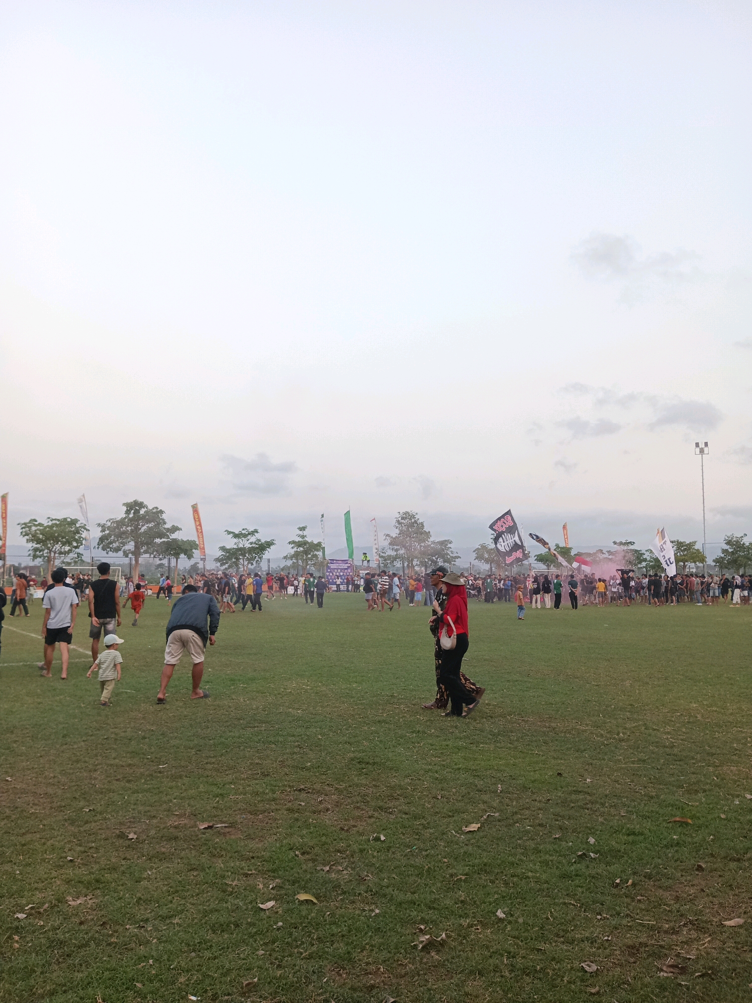 The image depicts an outdoor gathering on a grassy field, likely a sports field, at dusk. In the foreground, three individuals are visible: a child in a striped shirt and light-colored shorts, facing away from the camera and towards the right; an adult male in a dark blue shirt and light-colored shorts, bending over slightly and facing right; and a person in a red top and dark patterned skirt or pants, holding a white purse, standing and facing left.
The middle ground of the image is populated by a larger crowd of people, many standing and some appearing to be engaged in activities. Several flags are visible throughout the scene, including large black flags with white designs, and white flags with black lettering. One flag prominently displays what appears to be a stylized fish skeleton graphic. A plume of pink smoke is also visible rising from the crowd on the right side of the image.
In the background, trees line the edge of the field, and beyond them, some buildings and a cloudy sky are discernible. The overall atmosphere suggests a community event or festival taking place in Tawing, Indonesia, under an overcast sky as daylight fades. The presence of a soccer goal on the left side of the field further supports the idea of a sports-related or recreational event.