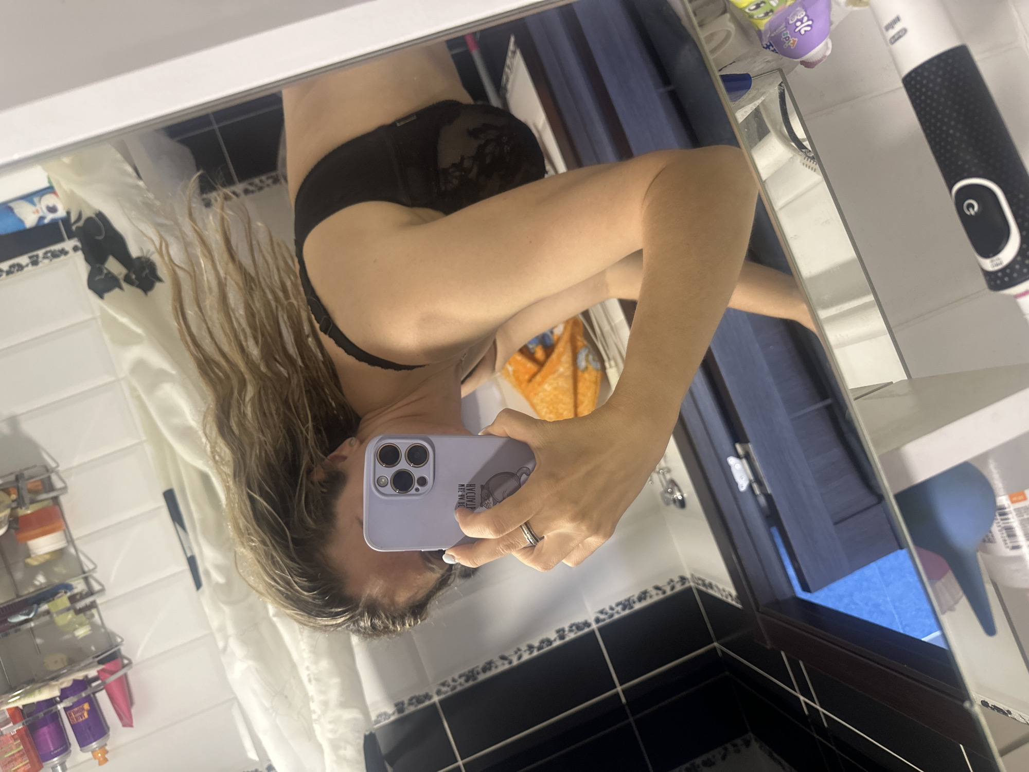 A woman is taking a mirror selfie in a bathroom. She is wearing a black bra and her hair is wet and down. She is holding a light purple phone with a cartoon image on the back. The bathroom appears to be in a residential setting. There are toiletries on a shelf in the background, and a shower curtain with a cat pattern. The woman is wearing a ring on her left hand. The image is taken from a medium distance. The lighting is artificial. The overall scene suggests a personal moment captured in a private space.