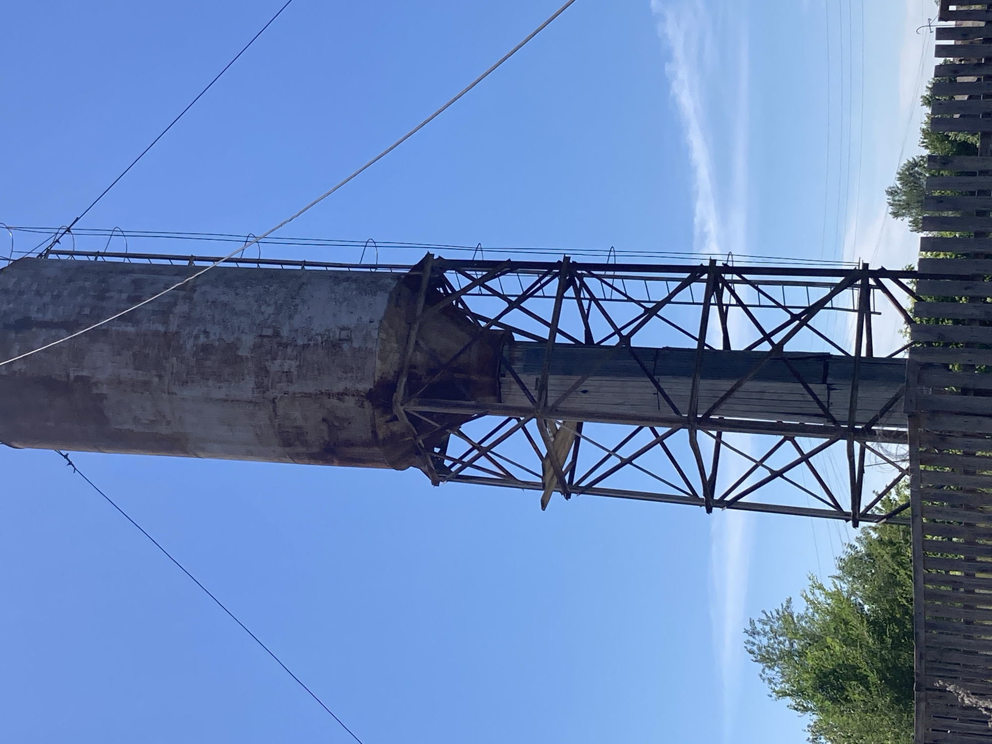An old, weathered water tower stands tall against a clear blue sky in the village of Rakity, Russia. The tower is cylindrical and made of concrete, showing signs of age with rust and discoloration. It is supported by a dark metal lattice structure. A ladder is attached to the side of the tower, providing access to the top. Several wires extend from the tower and are anchored to the ground, likely for stability. In the foreground, a rustic wooden fence runs horizontally, separating the area around the tower from a line of green trees behind it. The scene appears to be set during the daytime, with bright sunlight casting shadows. There are no people or animals visible in the image.