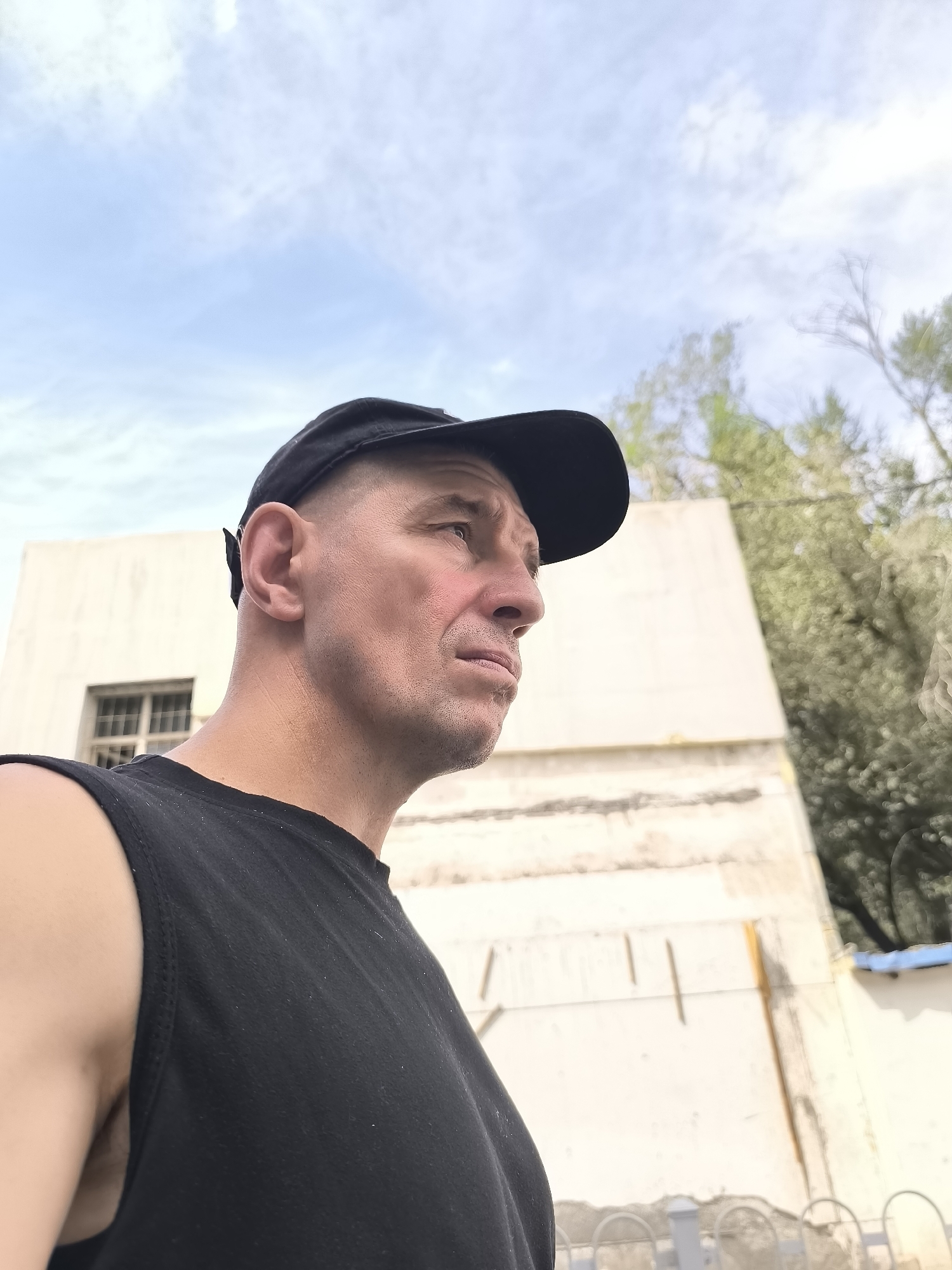A medium shot is captured from a low angle, featuring a Caucasian man in his late 40s or early 50s, wearing a black t-shirt and a black baseball cap. He is positioned on the left side of the frame, looking to the right with his profile visible. His skin is fair, and his hair is short and buzzed. In the background, a light-colored building with a window is visible, along with greenery and a pale blue sky with clouds. The overall impression is one of a candid outdoor shot, possibly taken in a residential or urban area. The man's gaze is directed towards something or someone outside the frame, suggesting a moment of contemplation or observation. The scene appears to be set in Ürümqi, China, given the provided context.