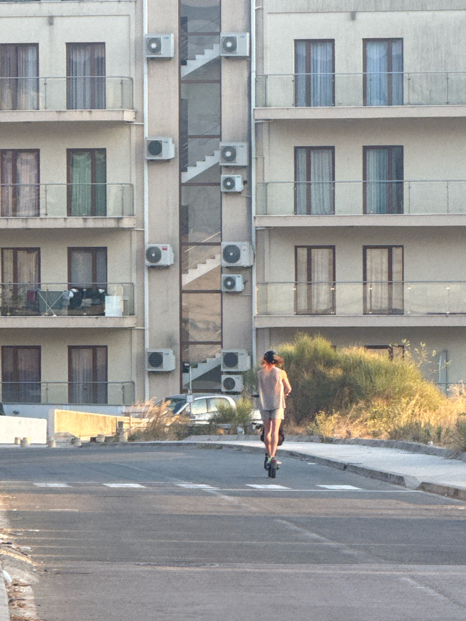 In Boreti, Montenegro, an individual on an electric scooter is captured from behind, traversing a road. The subject, wearing a light-colored sleeveless shirt and shorts, is positioned in the lower center of the frame, moving away from the viewer. The scooter appears to be dark in color with a visible rear wheel. The road in the foreground features a crosswalk with white markings. To the right of the road, a sidewalk and a section of dry, overgrown vegetation are visible, with a white vehicle partially obscured by the foliage. Behind the vegetation, a multi-story building with balconies and multiple windows is prominent. Air conditioning units are mounted on the exterior wall of the building. The architecture suggests a residential or hotel structure. The overall lighting indicates it is daytime, likely late afternoon or early evening, given the warm tones on the vegetation and building.