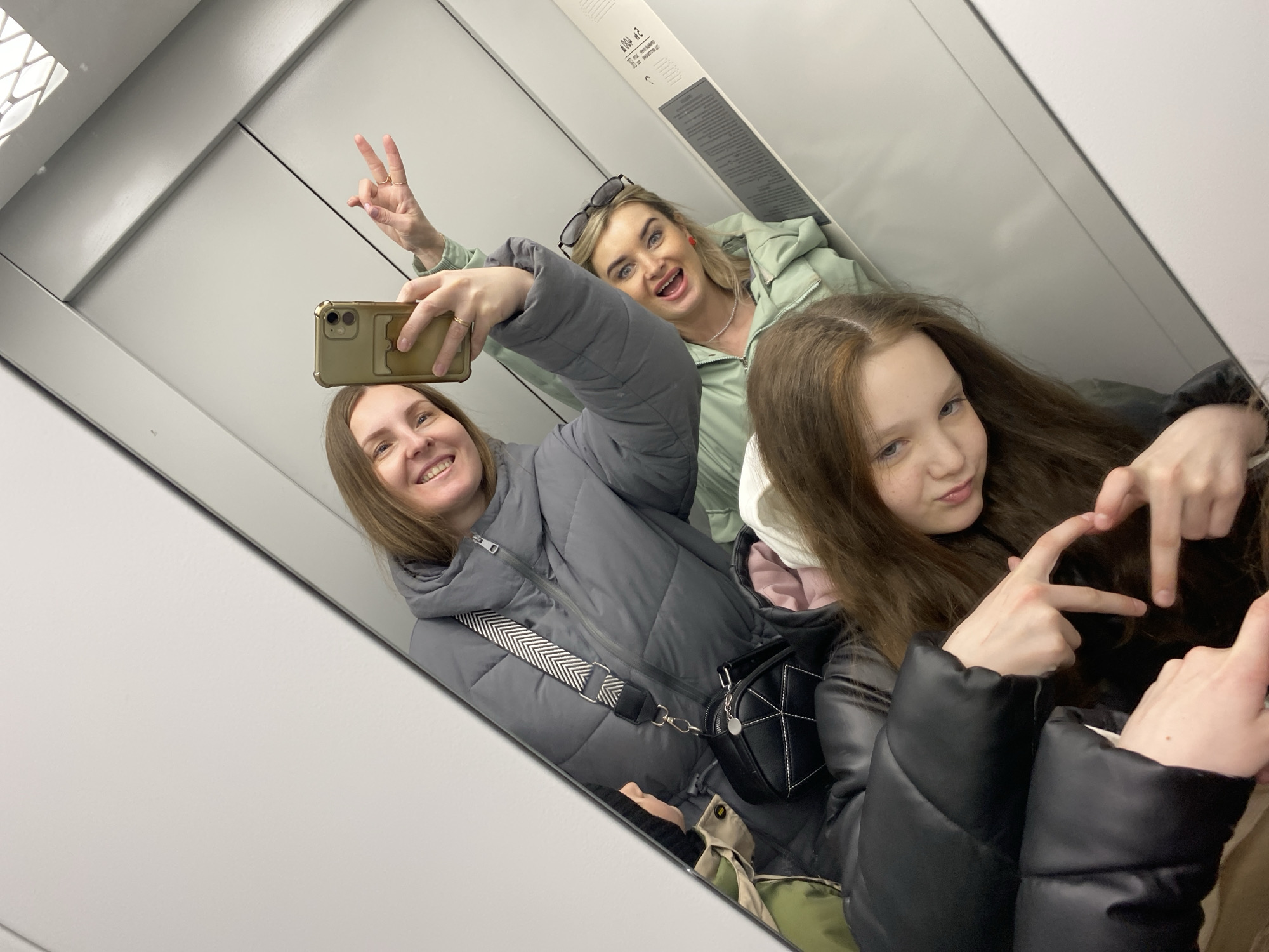 Three women pose for a selfie in an elevator, as reflected in the mirror. The elevator's interior is white and has a control panel. The woman taking the photo is wearing a gray puffer jacket with a black and white strap bag and is smiling. Behind her, another woman with sunglasses on her head is making a peace sign and sticking her tongue out. The third woman in the foreground is partially visible, making a heart shape with her fingers. The location is in Chaikovsky, Russia.