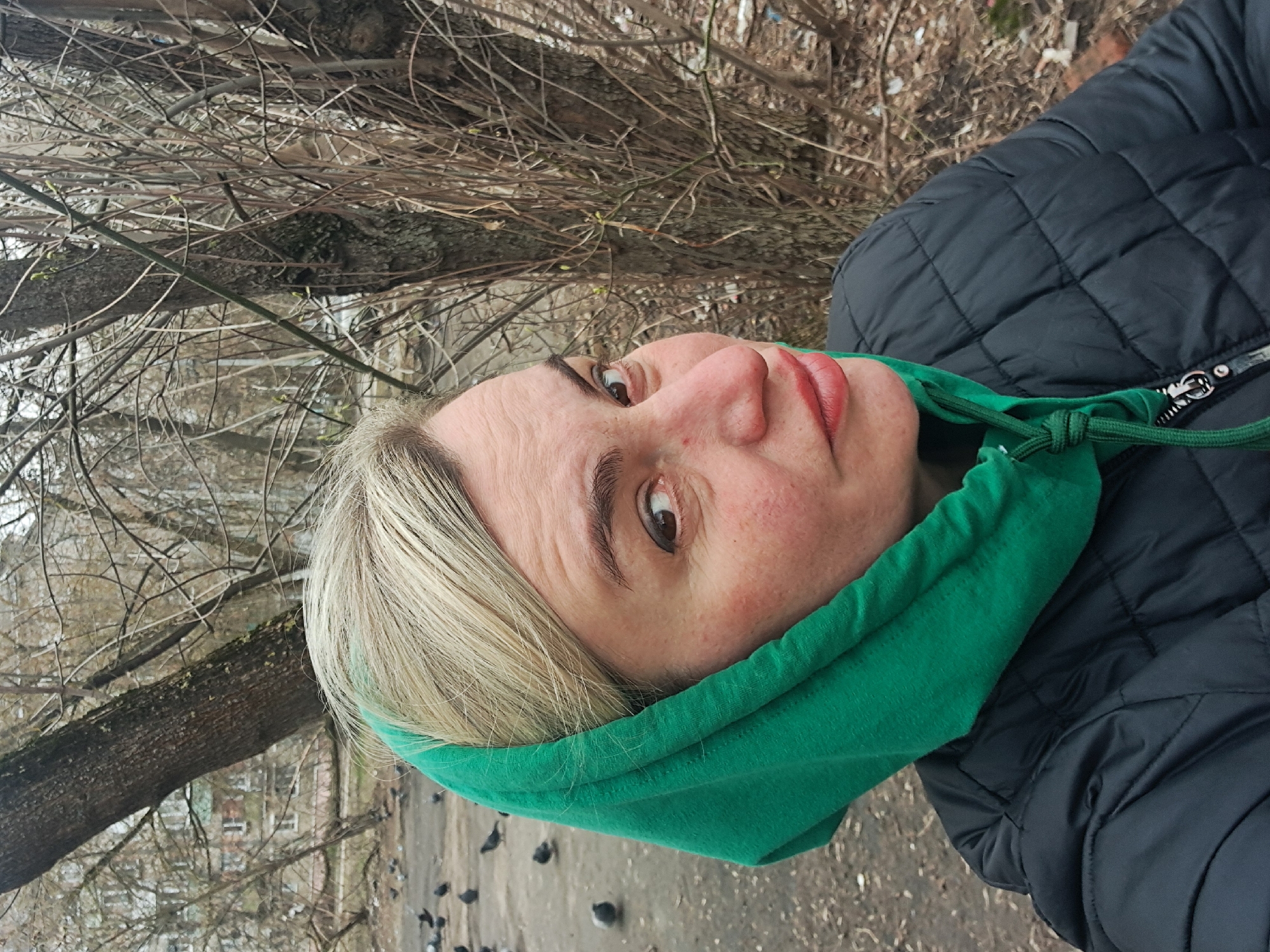A woman with blonde hair and wearing a green hoodie and black quilted jacket is standing outdoors. The background suggests a park or outdoor area in autumn or early spring, as indicated by the bare trees and visible branches. Pigeons are scattered on the ground. The location is Elektrostal, Russia, based on the buildings and overall environment visible in the background.