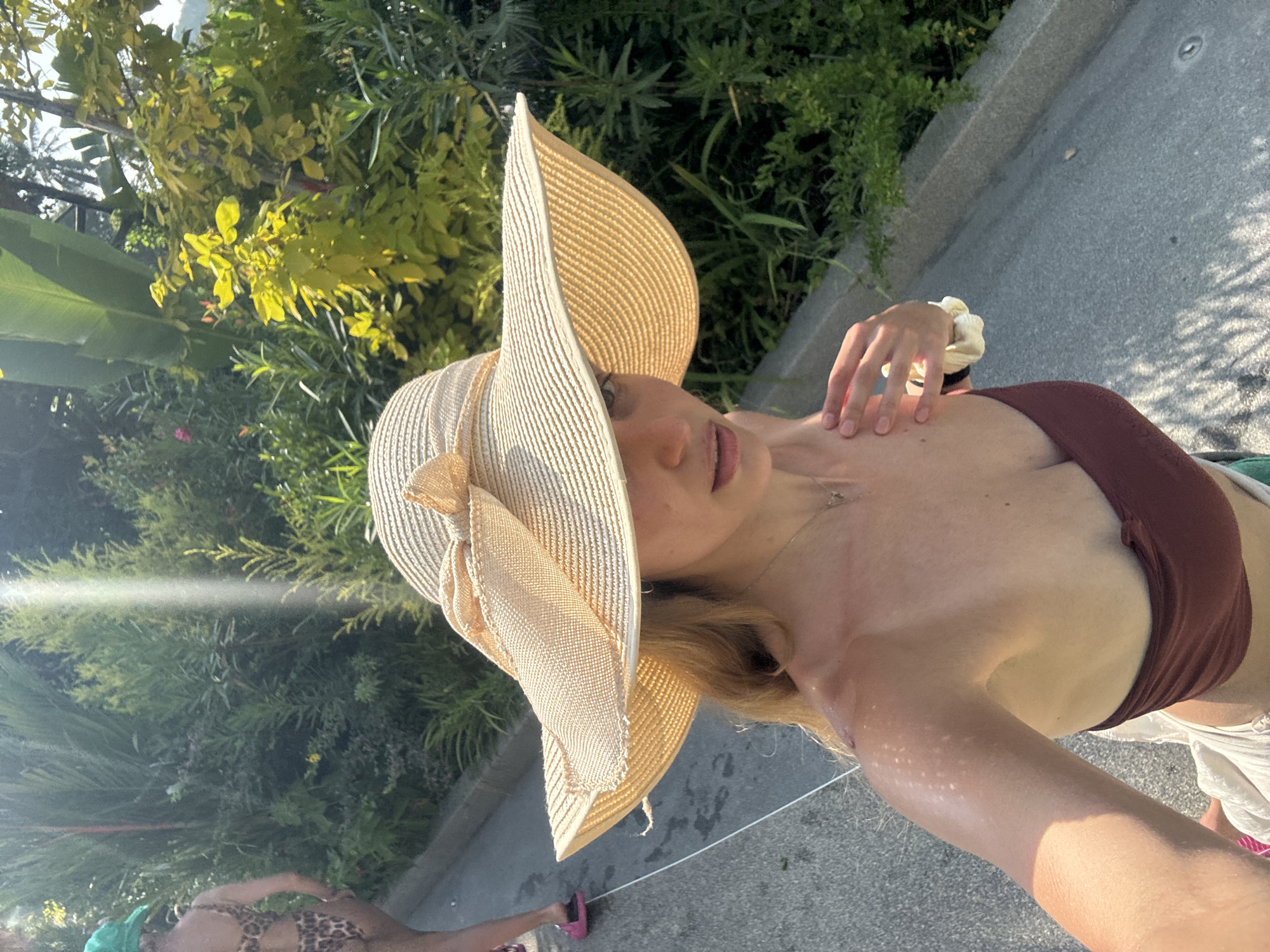 A woman wearing a wide-brimmed straw hat and a brown bikini top is standing outside in Ban Khlong Khian, Thailand. She has blonde hair and is wearing a necklace. Her right hand is resting on her left shoulder. The background consists of greenery and a paved area. A second person in leopard-print clothing is visible in the lower left corner of the image.