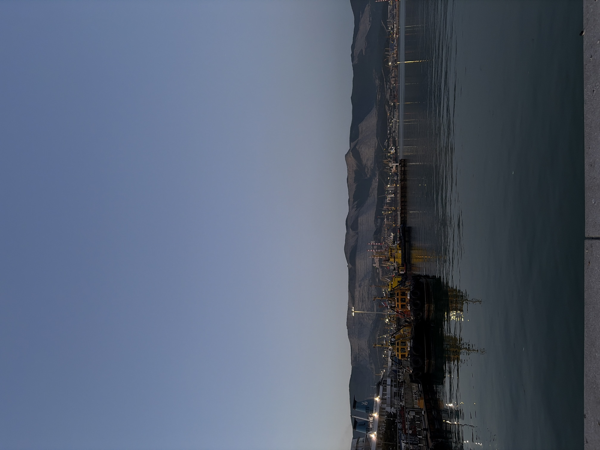 The image depicts a nighttime cityscape of Novorossiysk, Russia, situated along the coast. Mountains form a backdrop behind the city. The foreground shows dark water reflecting lights from the city, indicating a harbor or bay. The city itself is illuminated with numerous lights, suggesting an industrial or port area with docks and possibly ships. A concrete structure is partially visible on the right side of the image.