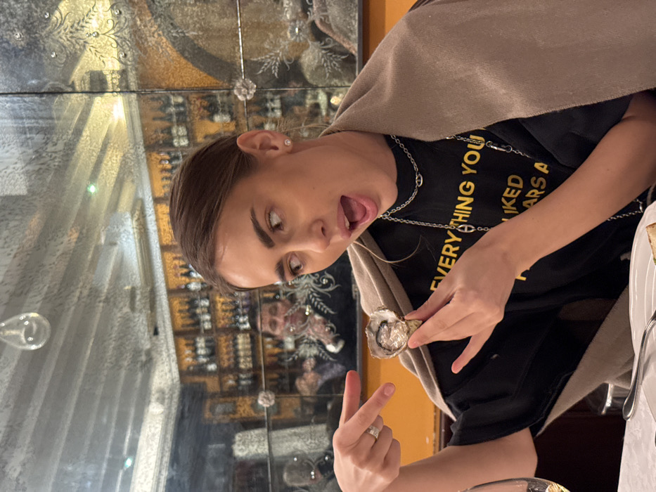 The image depicts a young woman seated at a white-clothed table in a restaurant located in Venice, Italy. She holds an open oyster in her right hand, pointing to it with her left index finger. Her mouth is open, and her gaze is directed off-camera to her left. She wears a black t-shirt with partial text "EVERYTHING YOU LIKED AARS A" visible, layered silver-tone chain necklaces with distinctive links, and a beige shawl draped over her shoulders. The background features a wall composed of large, etched, antique-style mirrored panels. Reflected prominently in these mirrors is a man in a dark, embroidered jacket, holding what appears to be a flute. Behind the man's reflection, golden shelves displaying numerous wine bottles are visible. A partial wine glass is on the table in the foreground.