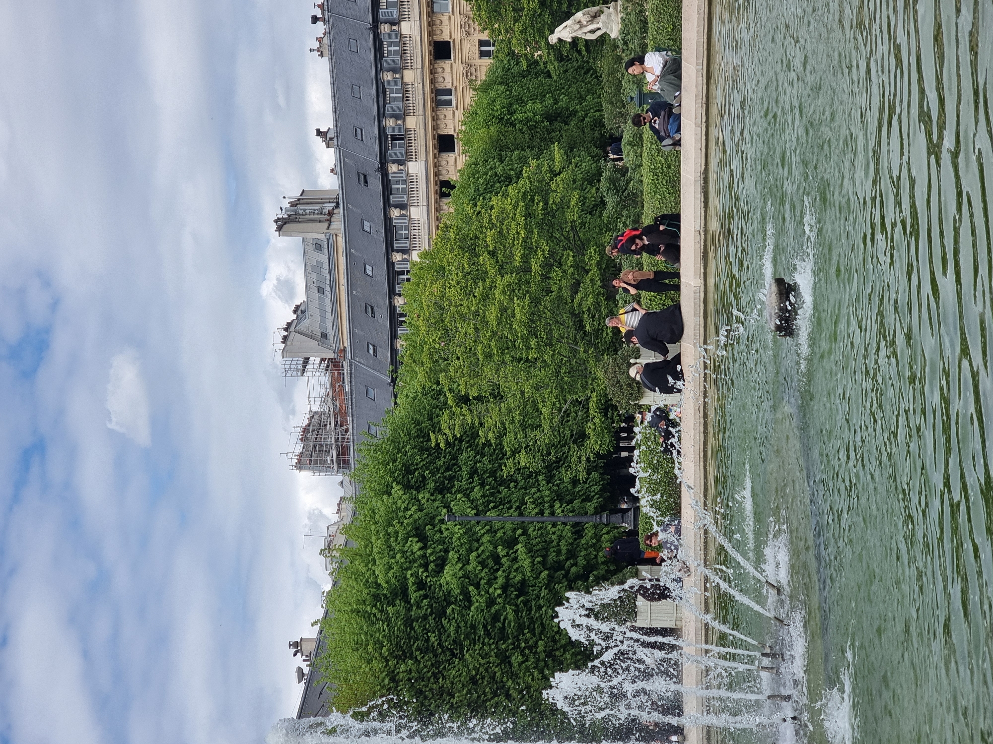 This image depicts a vibrant public park scene in Paris, France, on an overcast but bright day. The foreground is dominated by a large, rectangular fountain or pond with greenish water. Several powerful jets of water erupt from the surface, creating dynamic splashes and ripples across the water.
Along the stone edge of the fountain, numerous people are visible. They are casually dressed, some sitting on benches or the low retaining wall, while others stand, observing the fountain or simply enjoying the atmosphere. Among them, a person in a white top and dark pants is seated on the right, and another in a black top is seated further left.
Behind the people and a neatly trimmed hedge, a dense expanse of lush green trees forms a natural barrier, extending across the mid-ground. A classical white statue, depicting two figures, is partially visible nestled within these trees on the upper right side of the image. An ornate, dark lamppost also emerges from the foliage.
In the background, characteristic Parisian architecture fills the frame. Multi-story buildings with distinctive dark gray slate roofs, numerous windows, and prominent chimneys stretch across the horizon. One building features a more ornate, lighter-colored facade with balconies, while another displays scaffolding, indicating construction or renovation. The sky above is largely cloudy, with patches of lighter clouds. The scene suggests a tranquil moment of leisure in a bustling city park. No text is visible.