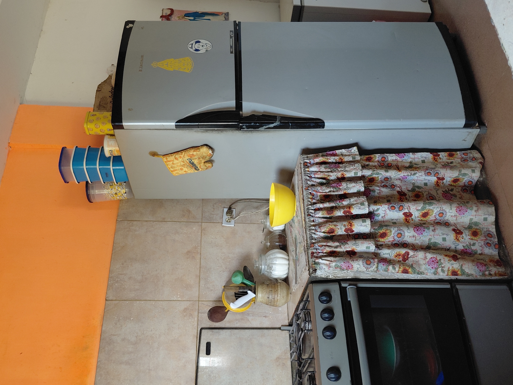 The image depicts a corner of a kitchen. The wall above the tiled backsplash is painted a vibrant orange. To the right, a tall, light gray refrigerator stands against a white wall. On top of the refrigerator, several blue plastic food containers are stacked, along with a yellow tin and a cardboard box. A yellow oven mitt hangs on the front of the refrigerator. To the left of the refrigerator, a stove is visible, with a floral-patterned fabric skirt covering the area beneath the countertop. The countertop itself holds a vase with cooking utensils, a clear glass jar filled with white powder, a smaller jar with brown contents, and a bright yellow bowl. An electrical outlet with a plug is mounted on the tiled wall behind the countertop. This scene is located in the city of Santa Ana, Venezuela.