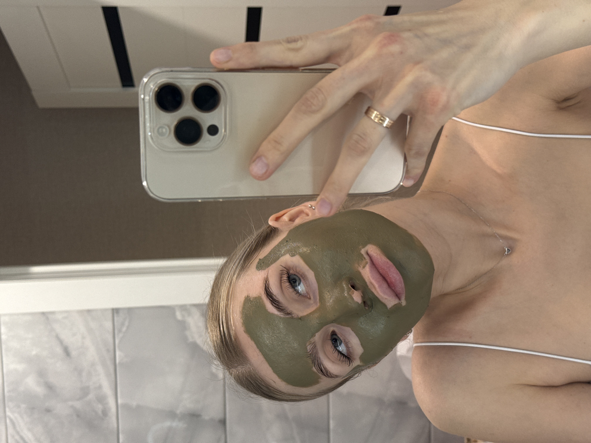 A fair-skinned woman is taking a selfie with her smartphone. She is wearing a green face mask, a thin-strapped white top, and a gold ring on her left ring finger. Her light-colored hair is pulled back. The phone is in a clear case with three camera lenses visible. The background includes a white vanity or cabinet and a tiled wall. The location is difficult to determine from the image, and there is no information to specifically confirm its location in Izhevsk, Russia.