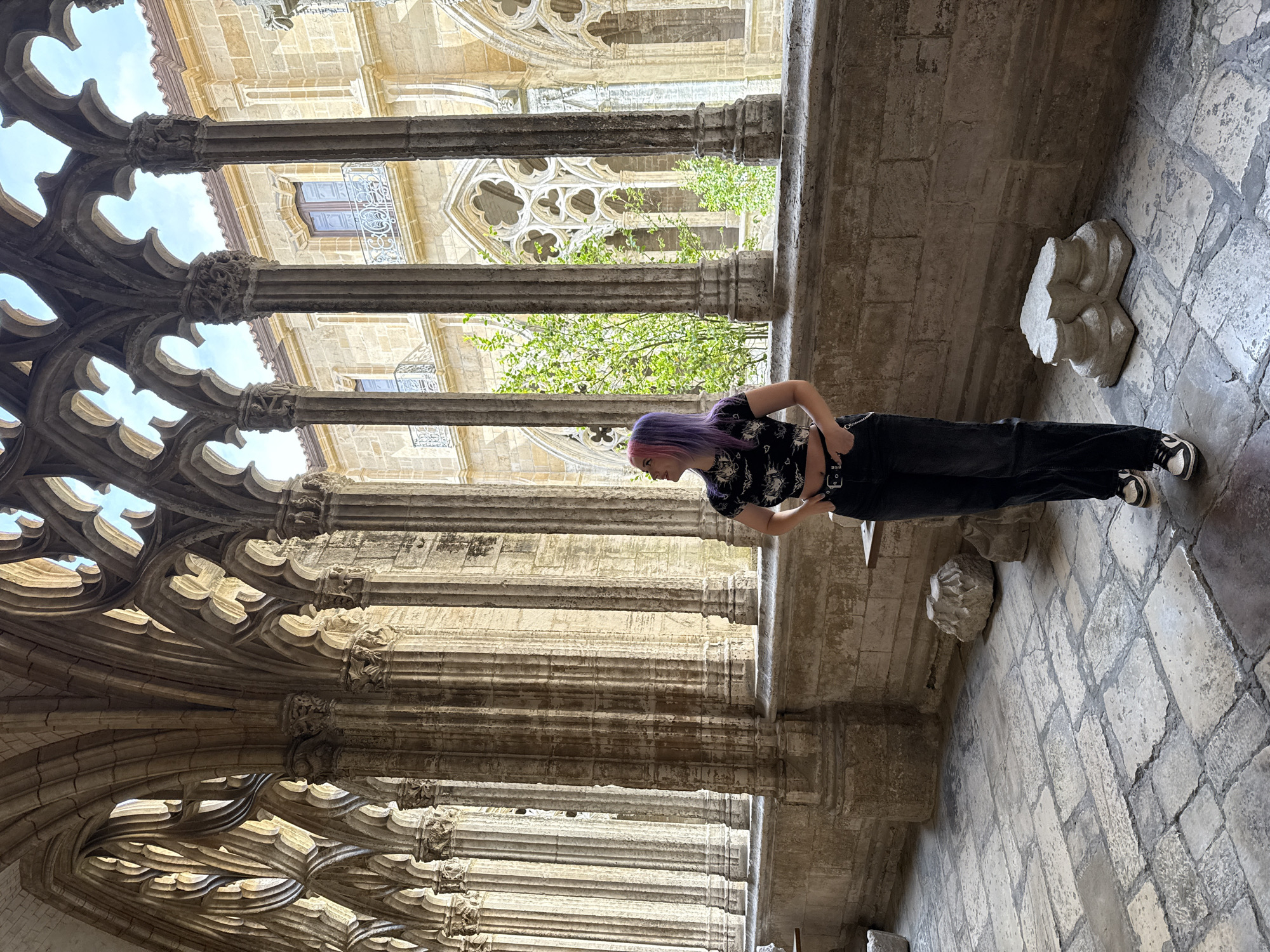 A person with purple hair, wearing a black patterned shirt and black pants, is standing in a stone structure. The structure is characterized by ornate Gothic-style arches and columns. The location is Unknown, Unknown. The person is positioned between the columns, leaning slightly forward. Behind the columns, there's a view of a building with windows and foliage. The ground is paved with stone blocks, and there's a stone object near the person's feet.
