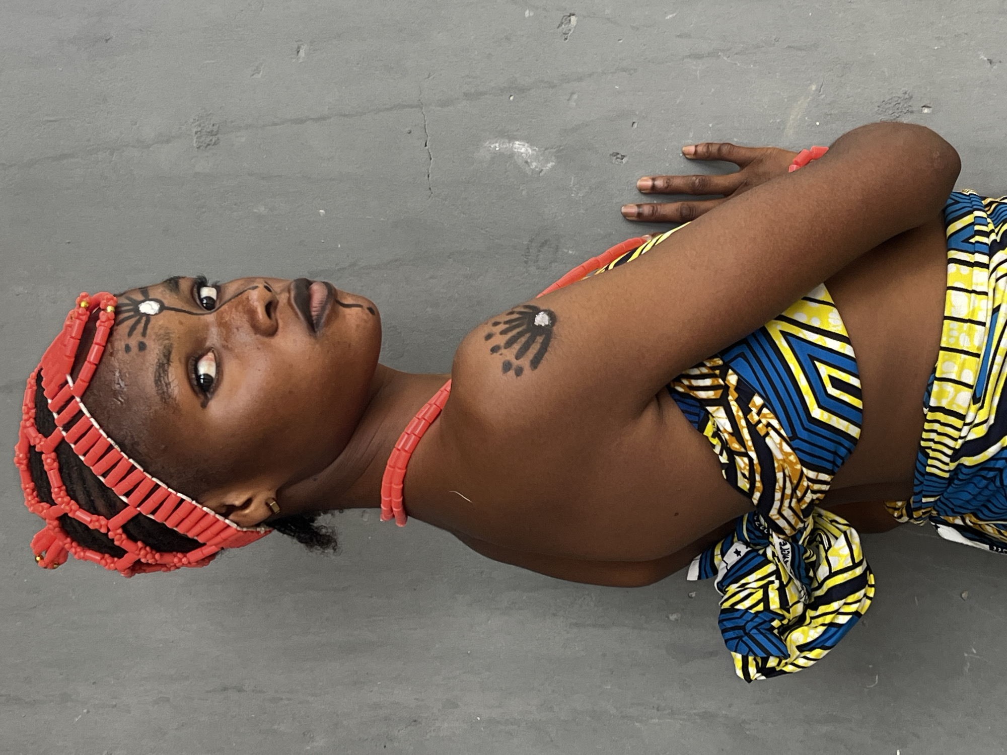 A woman lies on a gray surface, likely a floor, in an apparent artistic pose. She wears traditional Nigerian attire, including a headdress of red coral beads, a coral bead necklace, and a patterned cloth tied around her torso, exposing one shoulder. Facial markings in black are visible on her forehead and cheek. A black tattoo-like design is present on her upper arm. Her arms are crossed, with one hand resting near her head and the other near her side. The location, Badagry, Nigeria, is not directly discernible from the image itself; however, the traditional clothing and adornments suggest a cultural context within Nigeria.