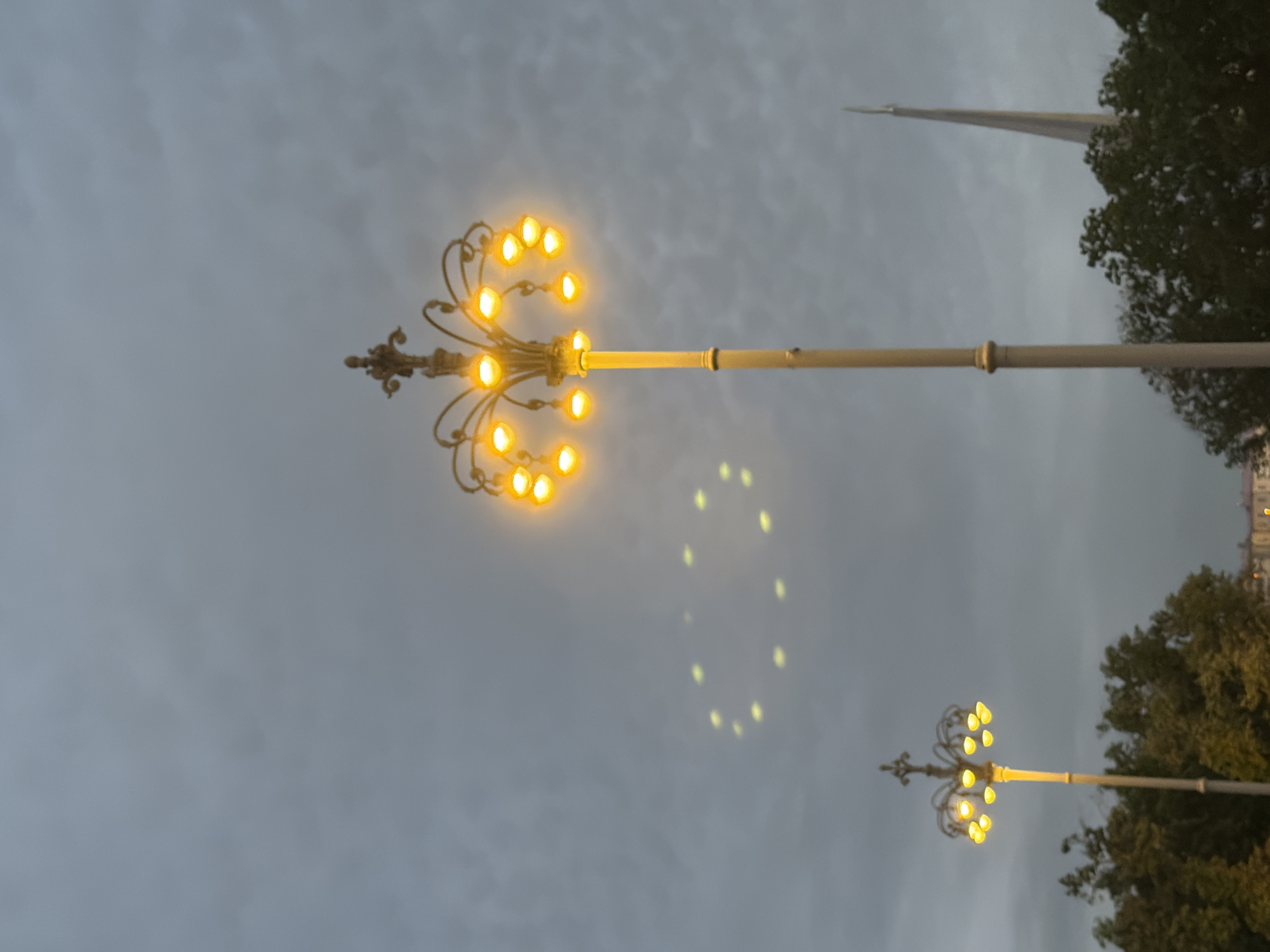 Two ornate lampposts are illuminated against a cloudy, gray sky. The lamppost on the right is taller and has a more elaborate design with multiple lights arranged in a circular pattern. The lamppost on the left is shorter and has a simpler design with fewer lights. A shadow of the lights from the right lamppost appears as a ring of glowing dots in the sky. In the background, dark green trees line the bottom of the frame, and a tall, pointed structure, possibly a spire, is visible on the far right. The overall scene suggests an outdoor public space at dusk or during overcast weather.