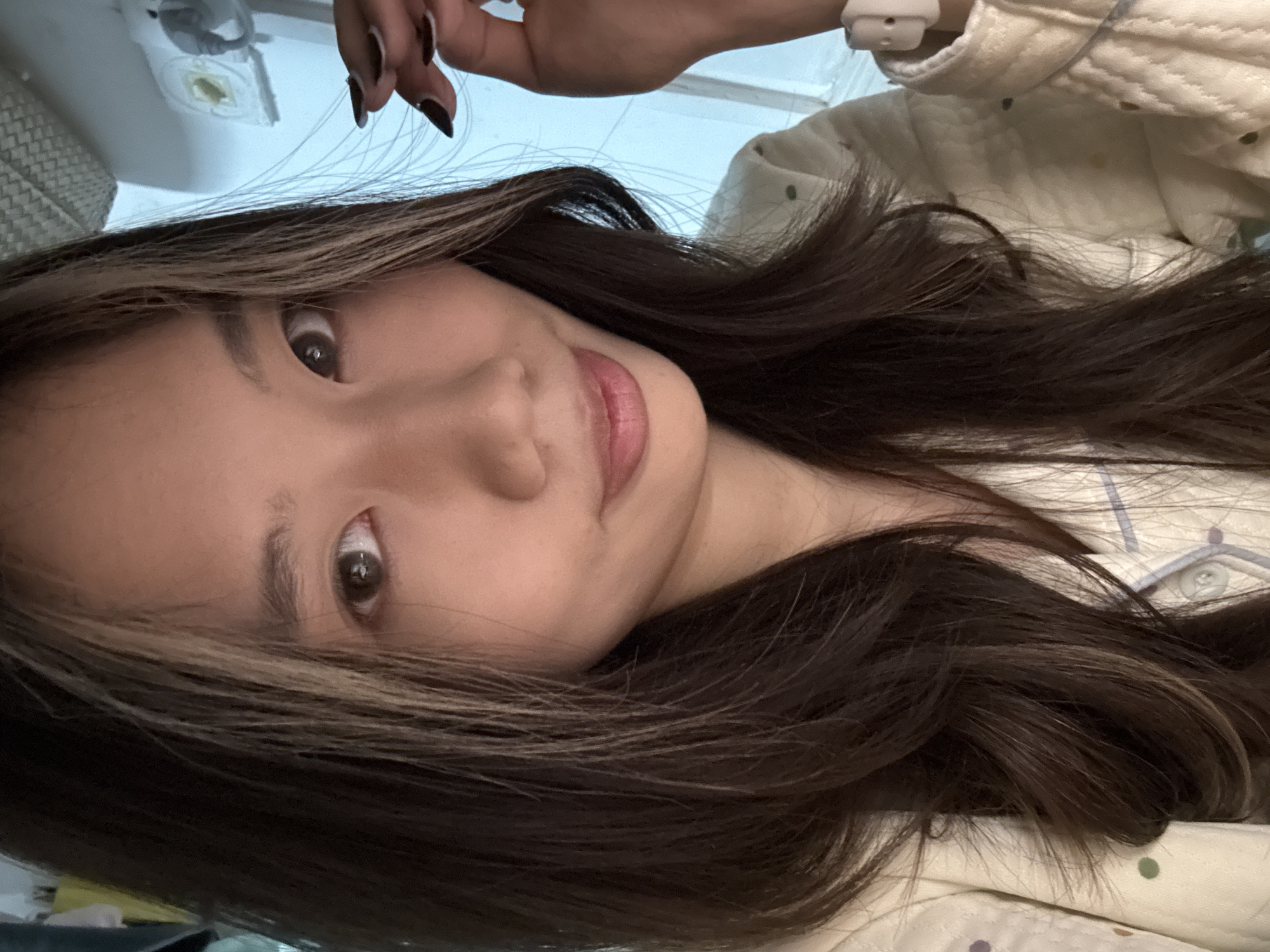 A close-up portrait depicts a young adult female, identified as East Asian, situated indoors in what appears to be a private dwelling in the city of Nan'an District, China. The subject faces the camera directly, displaying dark brown eyes and a subtle smile with slightly parted, pink lips. Her long, dark brown hair features lighter, possibly blonde, highlights, particularly framing the right side of her face. Her right hand is raised to eye level, fingers slightly curled, with fingernails painted black. A white wristband is visible on her right wrist. She is wearing a light-colored, possibly cream, quilted top with small, multi-colored polka dots. The background consists of indistinct indoor elements, including a light blue surface to the right, adjacent to white architectural elements suggestive of a window frame or wall fixtures, one being a white rectangular object and another with a circular yellow center. No specific identifying landmarks for Nan'an District are visible.