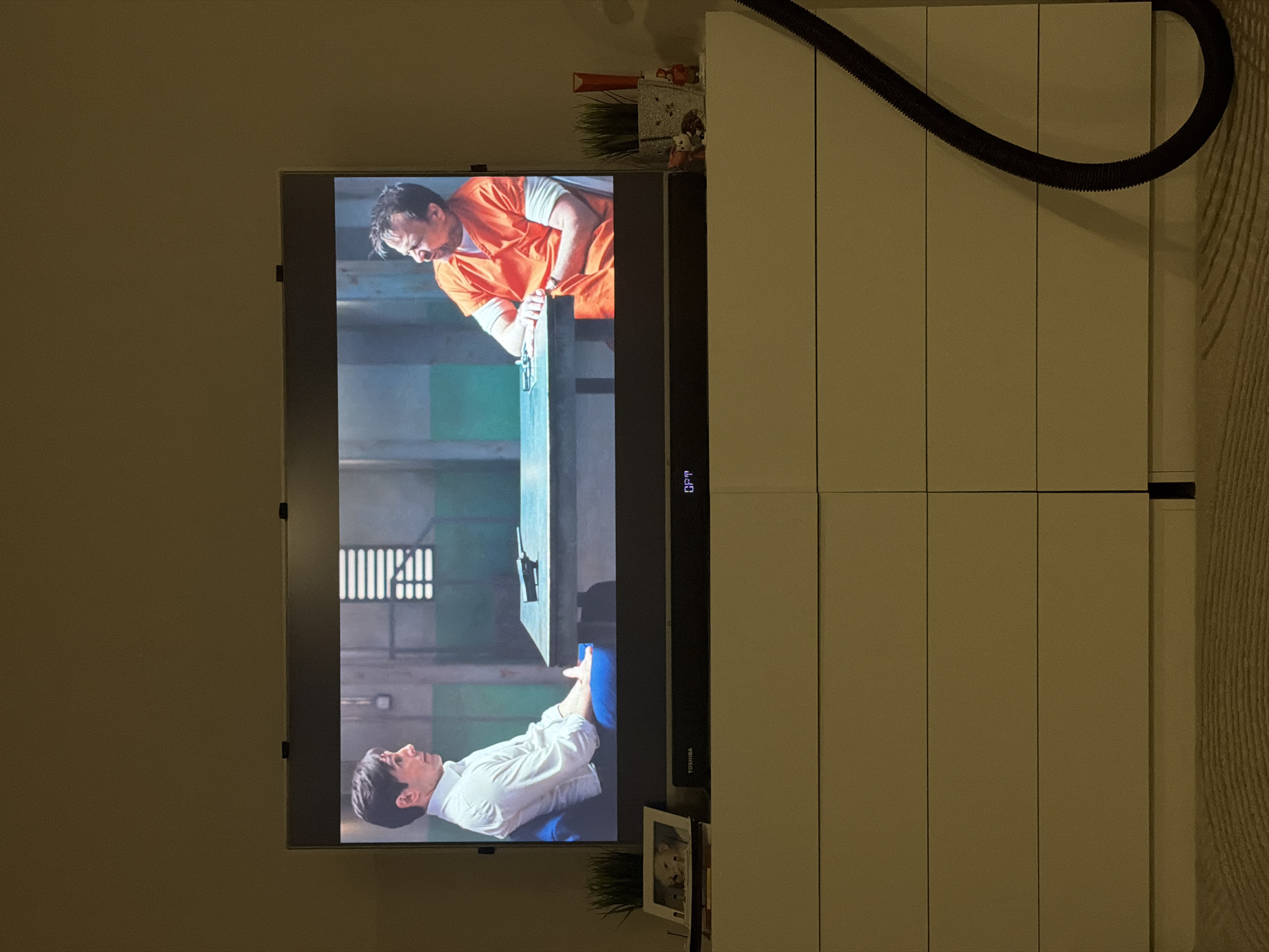 A television is mounted on a wall, displaying a scene from what appears to be a film or show. On the screen, two men are featured: one dressed in an orange jumpsuit and the other in a white button-down shirt. The setting is a stark, industrial environment with what looks like a prison interior, including barred windows. Below the television, there's a decorative item, potentially a framed photo or small plant. To the right, there is a tall, white cabinet with vertically aligned doors. A black hose-like object runs along the top of the cabinet. The background wall is a solid, neutral color. The environment around the objects in the room is fairly basic and appears to be an interior setting. There is no information available to determine if the location is in Chaikovsky, Russia.