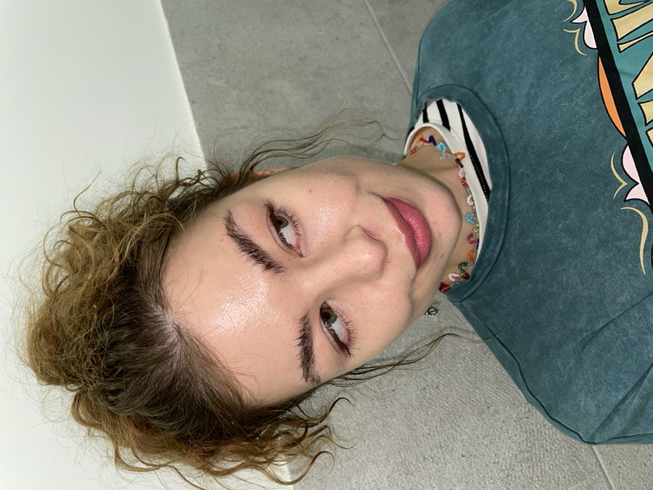 The image displays a close-up, slightly angled portrait of a single female individual against an indoor background. The background features a light grey wall in the lower section, characterized by a horizontal seam, and a plain white wall in the upper section. The specific location within the city of Budva, Montenegro, is not discernible from the visual evidence presented.
The subject is an adult female with light brown, curly hair pulled up into a messy bun. Her skin appears light-toned with a visible sheen on her forehead and cheeks. She has dark, thick eyebrows styled upwards and hazel eyes directed towards the viewer's left. Her lips are closed, exhibiting a reddish-pink tint, and form a subtle smile.
She is dressed in a white and black horizontally striped shirt, worn underneath a teal or dark green long-sleeved crewneck sweatshirt. A colorful graphic design, incorporating orange, light blue, and pink elements, is partially visible on the front of the sweatshirt. She wears a multi-colored beaded necklace around her neck, composed of small, distinct segments of various hues. A small, dark-colored earring is visible on her left earlobe.