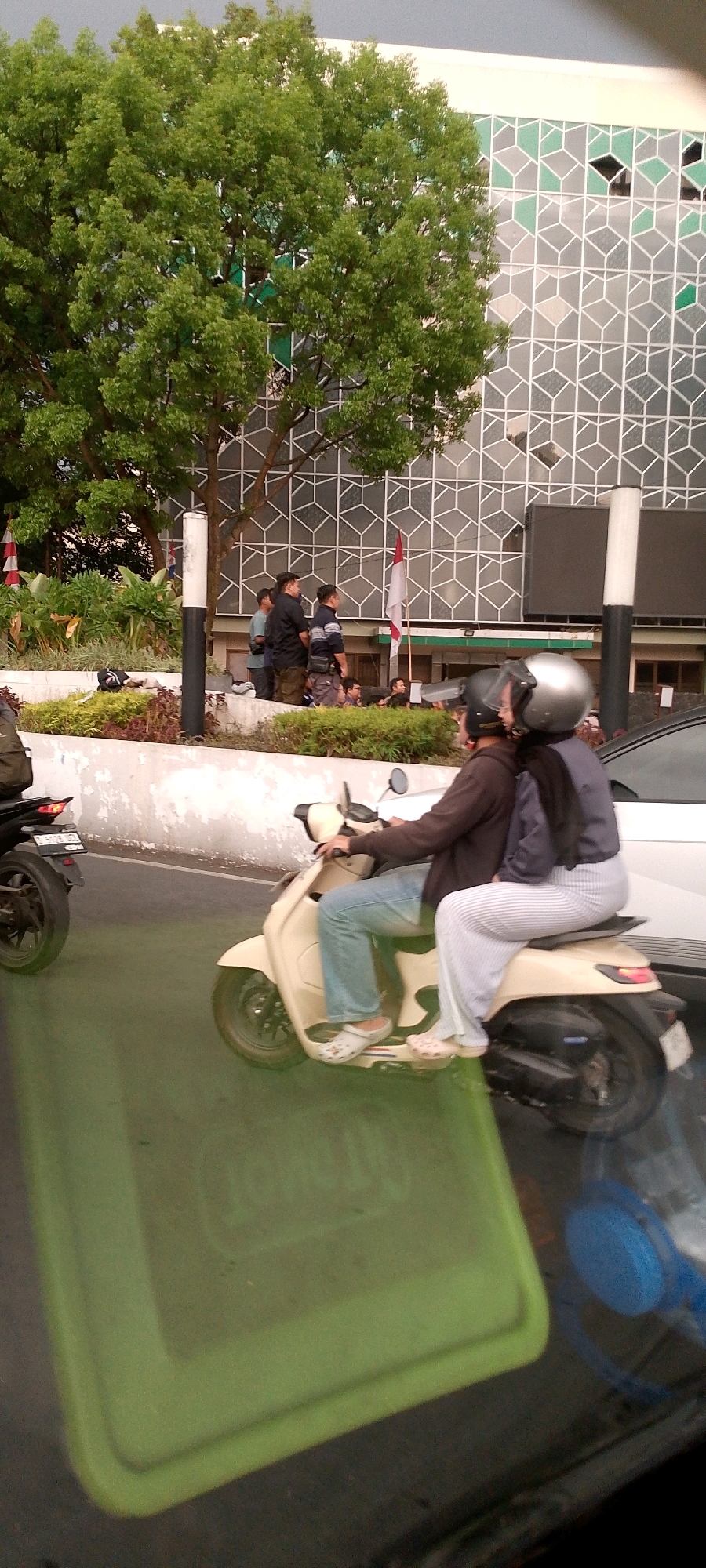 The image depicts a street scene in what appears to be Bandung City, Indonesia. A cream-colored scooter is in motion, carrying two individuals. The rider, a male, wears a brown jacket, blue jeans, and white clogs. The passenger, a female, is seated behind him, wearing a dark hijab and striped pants. She also wears a silver helmet.
In the background, a large tree with lush green foliage is visible, partially obscuring a modern building with a distinctive geometric facade composed of white and green hexagonal patterns. Several people are standing near a low wall in the mid-ground, including men in dark clothing and what appear to be security personnel. A red and white Indonesian flag is planted near the building. The foreground is dominated by a bright green rectangular object, possibly a container or a piece of equipment, and the dark, reflective surface of what could be a vehicle's windshield or a glass table. The road surface is partially covered by a green layer, possibly a reflective lane marking or a green mat.