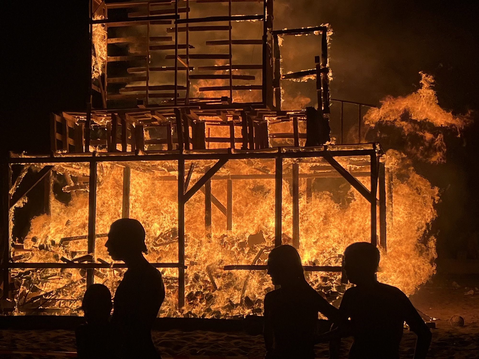 In the city of La Línea de la Concepción, Spain, a large wooden structure is engulfed in flames at night. The fire is intense, with bright orange flames consuming the wooden beams and planks of the structure, which appears to be multi-leveled and somewhat abstract in its design. Smoke billows upwards and to the right, obscuring the night sky. In the foreground, silhouetted against the fire, are at least three people. One person, appearing to be an adult, is standing and looking towards the fire, with another smaller figure, possibly a child, in front of them. To the right, another silhouetted figure stands with their back to the viewer, also facing the fire. The ground appears to be sandy or a beach. A dark, spherical object, possibly a ball, is visible on the ground to the far right. The overall scene depicts a controlled burning event, likely a bonfire or a ceremonial fire, with spectators observing the spectacle.