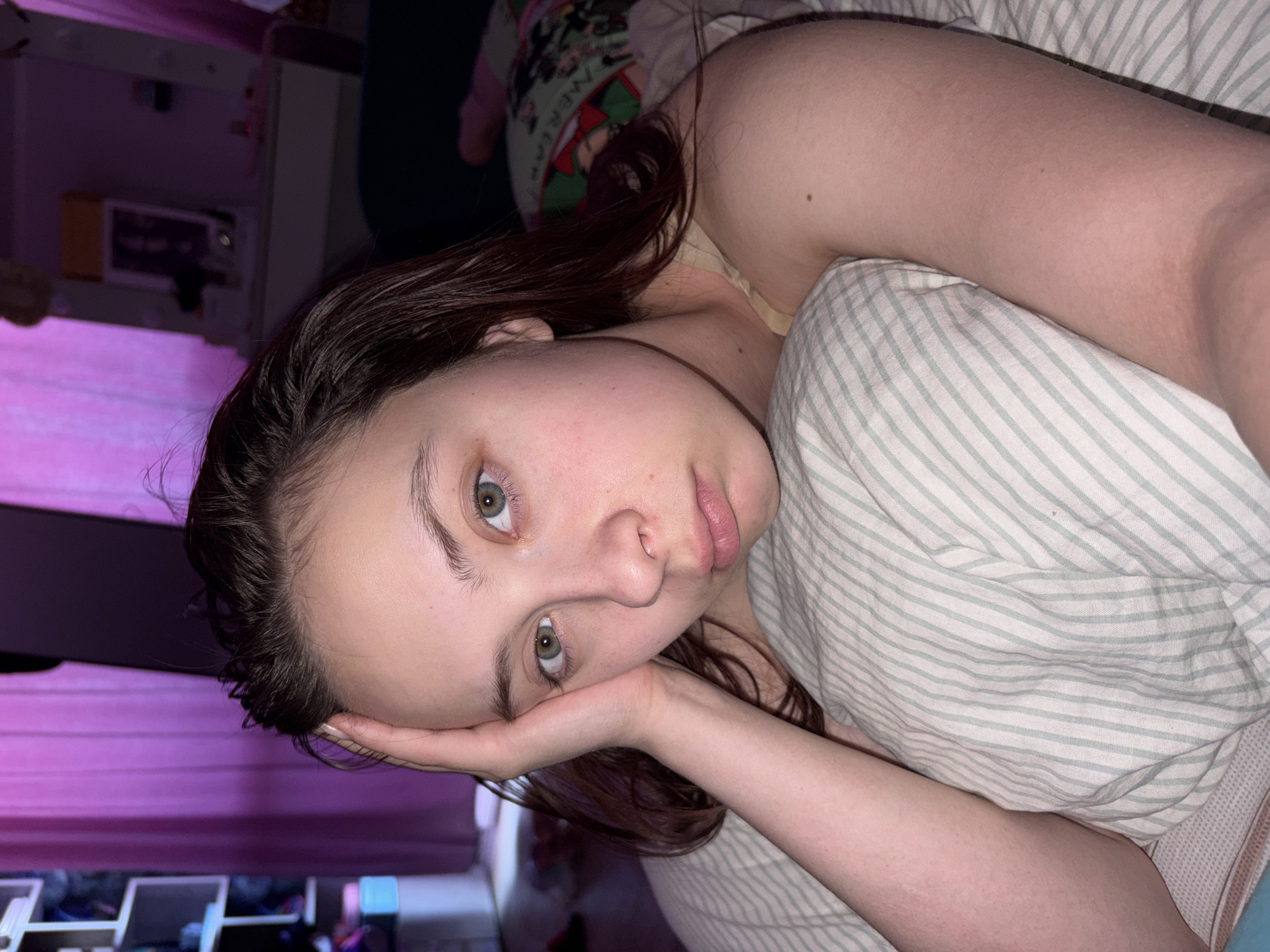 A young adult woman with fair skin and shoulder-length brown hair is lying down, resting her head on her hand. She has light green eyes and a neutral expression. She is on a bed covered with a white and light green striped sheet. The background includes a purple-lit wall or piece of furniture and some other objects that are out of focus. The image provides no information about Прикамский, Russia.
