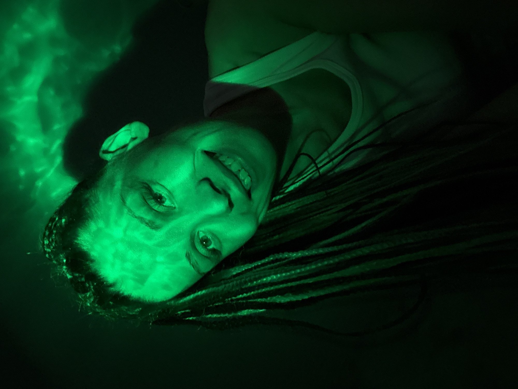 A woman with braided hair is illuminated by green light in an otherwise dark setting. She wears a white tank top and is smiling, looking towards the viewer. The background is dark, with some abstract patterns of green light. There is no information present in the image that would allow determining a location in the city of Rusanovo, Russia.