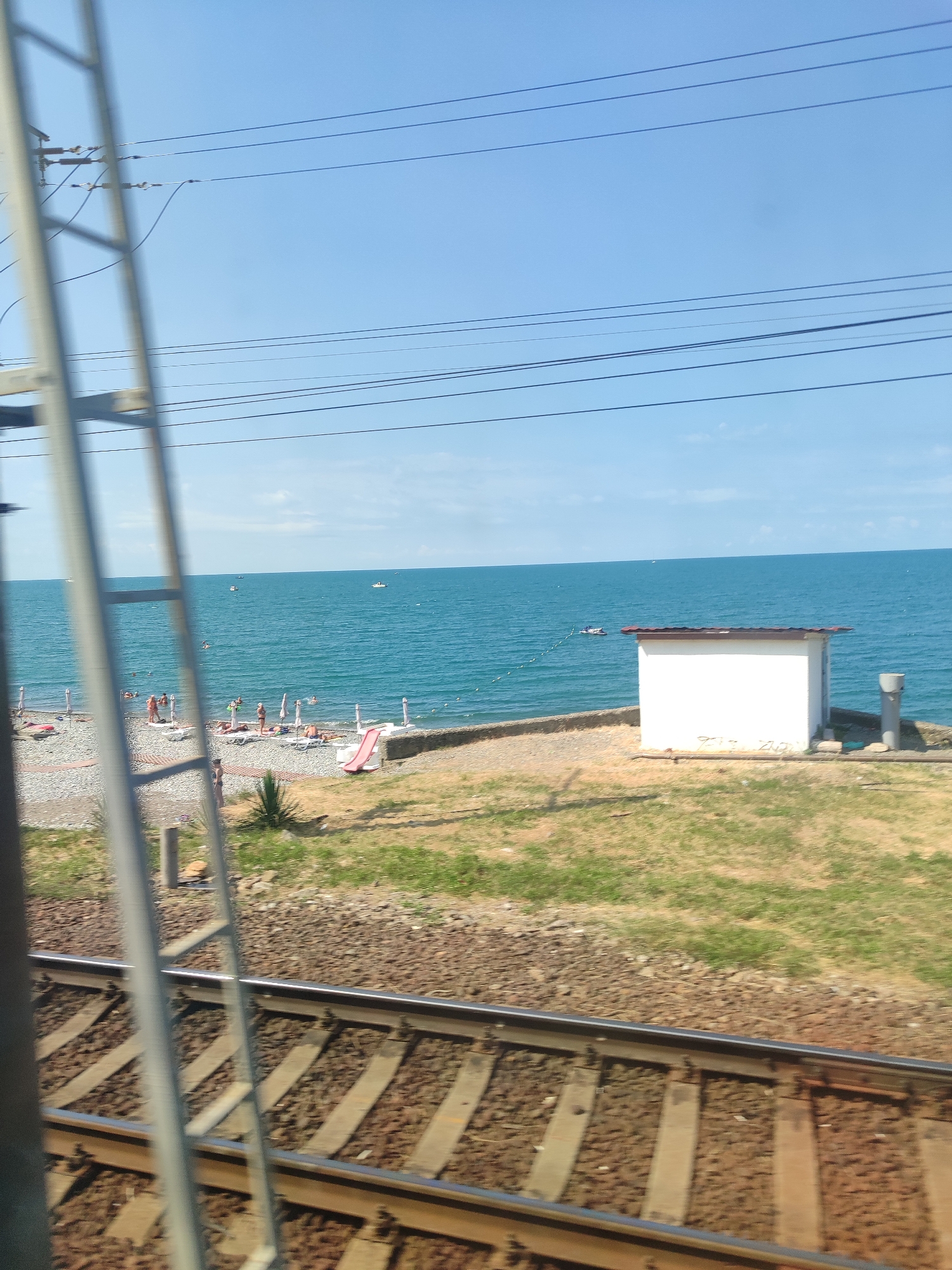 This media file captures a sunny day at a beach in Sochi, Russia, as viewed from a moving train. The foreground is dominated by the blur of train tracks, with a portion of the train's structure, possibly a ladder or part of the carriage, visible on the left.
Beyond the tracks lies a pebble beach populated with a few sunbathers lounging on chairs and towels. Some people are standing or walking along the shore. A red slide is set up on the beach, and several beach umbrellas provide shade. The turquoise sea stretches out to the horizon, where a few small boats are visible. In the middle distance, a small, white, shed-like building with a reddish roof sits near the water's edge. The sky is a clear, bright blue, indicating fair weather and likely midday. The overall scene evokes a relaxed, summery atmosphere.