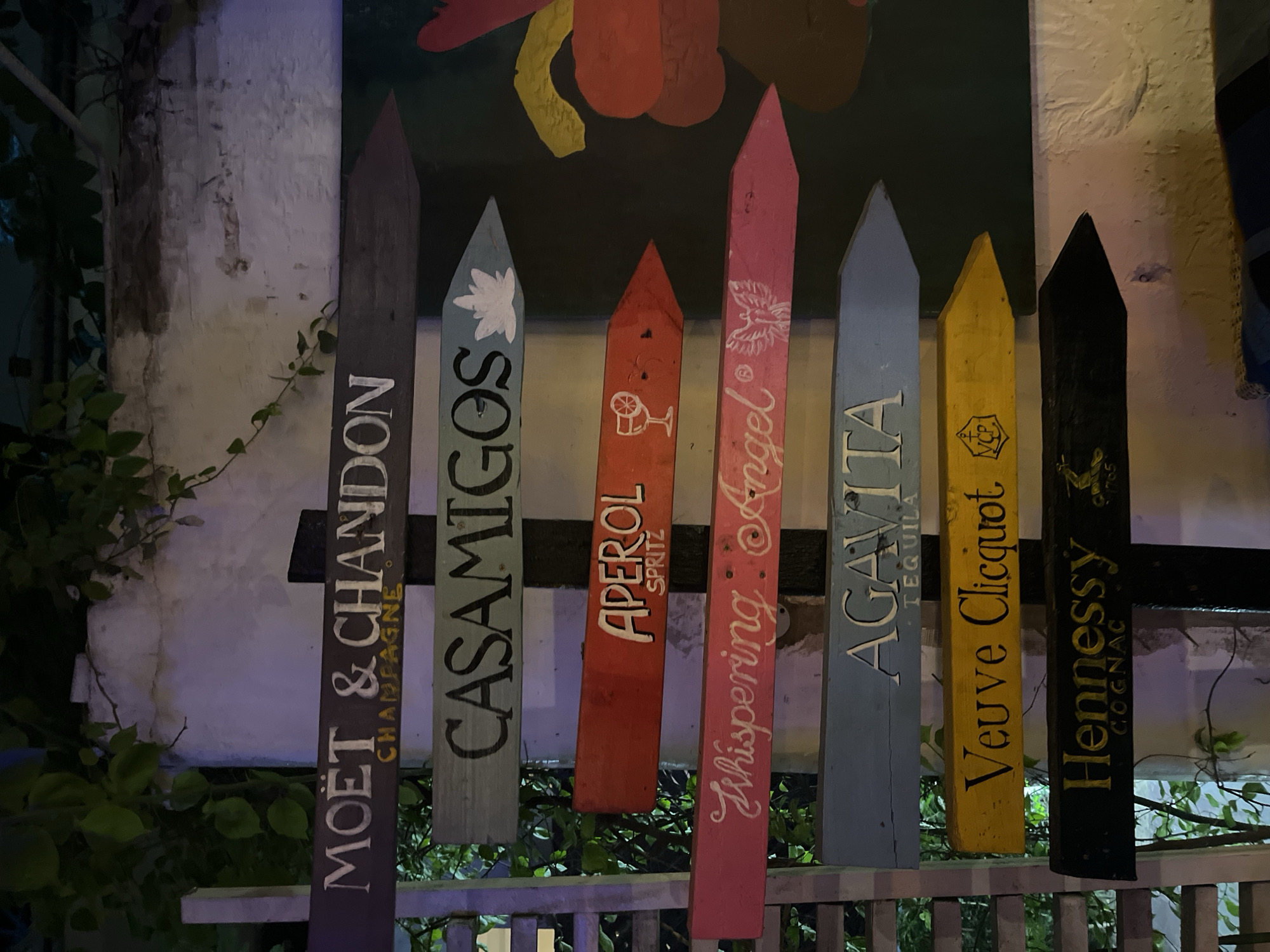 The image shows a directional signpost with multiple wooden planks, each pointing to a different brand of alcohol, suggesting a bar or a similar establishment. The planks are painted in various colors and display brand names like "MOËT & CHANDON CHAMPAGNE," "CASAMIGOS," "APEROL SPRITZ," "Whispering Angel," "AGAVITA TEQUILA," "Veuve Clicquot," and "Hennessy COGNAC." The signpost is set against a textured wall with some greenery, possibly vines, growing on it. The lighting suggests it is nighttime or a dimly lit indoor space. The overall scene is consistent with a themed bar, possibly in a tourist area. While the specific location in Accra, Ghana, cannot be definitively determined from the image alone, such a display is common in entertainment districts or upscale establishments.