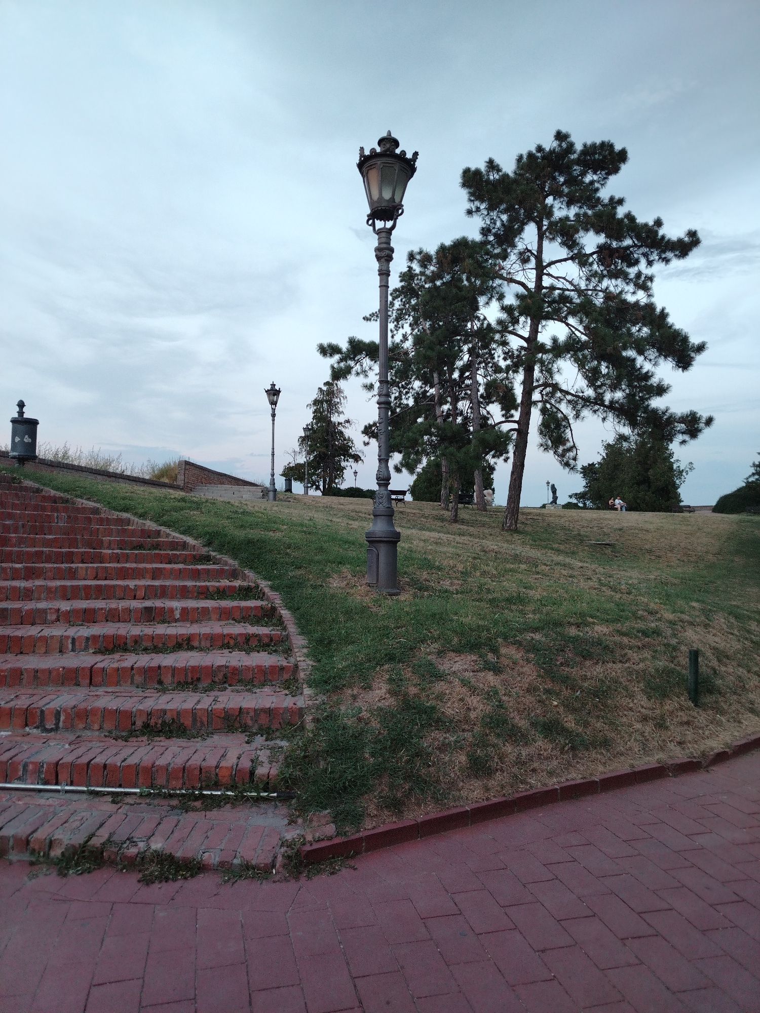 The image shows a view of Petrovaradin Fortress in Petrovaradin, Serbia, at dusk. In the foreground, a red brick staircase ascends a grassy slope. To the left of the stairs, there is a dark trash receptacle. A large, ornate lamppost stands prominently on the grassy area, with a second lamppost visible further in the background. Several trees, including pine trees, dot the landscape. In the middle ground, there are a few people sitting on a grassy area, and further back, a statue is visible. The sky is overcast and appears to be a pale blue-gray. The ground is paved with red bricks in the lower right corner.