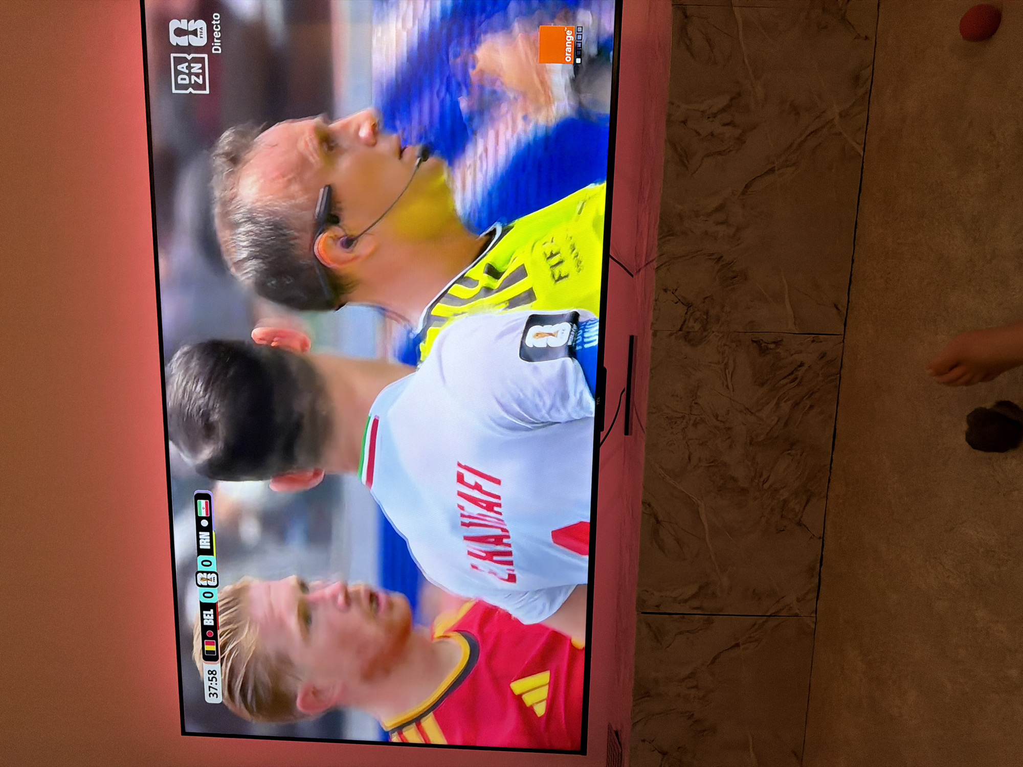 The image is a close-up shot of a television screen displaying a soccer match. The scoreboard at the top of the screen shows the time as 37:58 and indicates the score between BEL and IRN as 0-0. Two soccer players are visible on screen, one in a red and yellow jersey and another in a white jersey with red lettering. A referee in a yellow jersey with a microphone headset is also visible, facing right. The DAZN and FIFA logos are visible on the screen. The background of the image appears to be a dimly lit room with ambient lighting. There is no indication of the city of Barcelona or Spain within the image.
