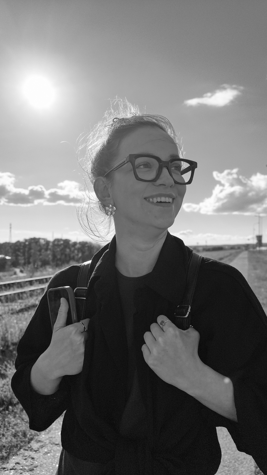 In a black and white, waist-up shot, a young woman with her hair pulled back smiles and looks to the right. She wears black-rimmed glasses, a dark jacket over a darker shirt, and a flower-shaped earring with sparkling stones. Her hands are positioned in front of her, one holding a smartphone and the other resting on her jacket. Rings adorn two fingers. The background shows a bright sun in the sky with scattered clouds, railroad tracks, and trees on a slightly hazy horizon. The scene appears to be outdoors, possibly near a railway line.