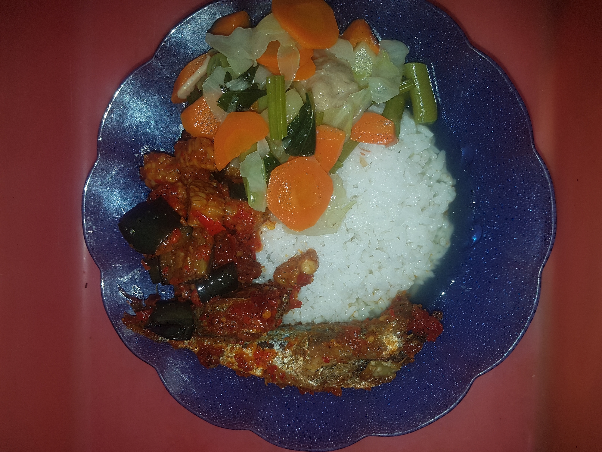 A close-up, top-down shot shows a blue, scalloped plate filled with Indonesian cuisine. The plate sits on a red surface. White rice forms the base of the meal, occupying the center and right portion of the plate. To the left of the rice is a whole fried fish coated in a thick, red chili sauce. Next to the fish and rice are cubes of fried tempeh and slices of fried eggplant, also coated in a similar red sauce. To the right of the rice and vegetables are mixed stir-fried vegetables including sliced carrots, cabbage, and green beans. The image does not provide information about a specific location within the city of Tinatar, Indonesia.
