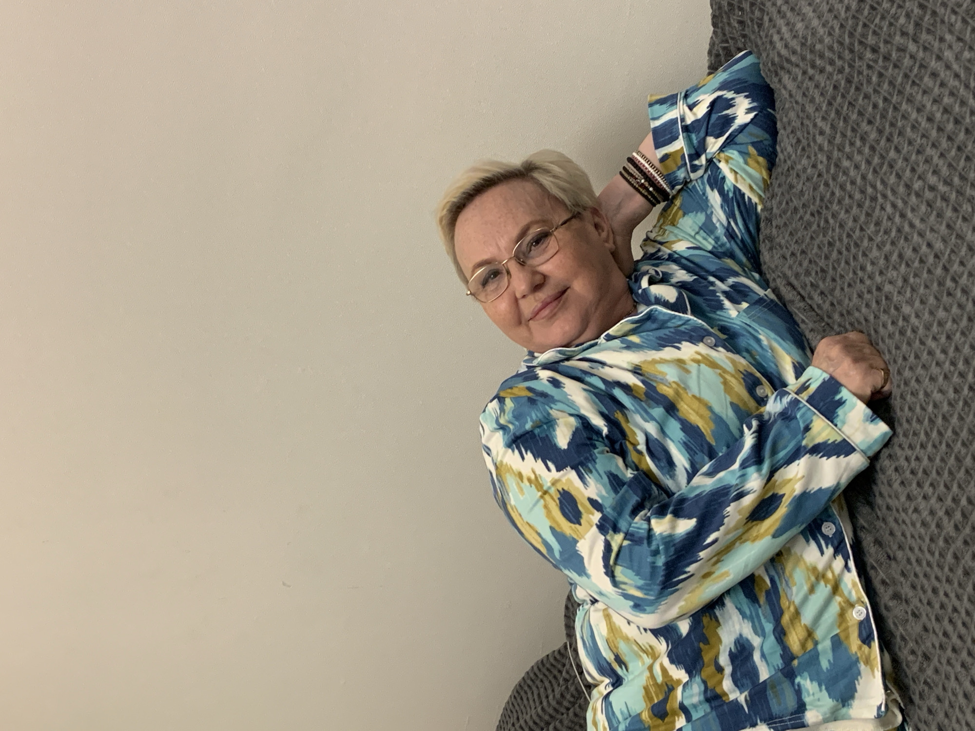 A woman, appearing middle-aged, with short blonde hair and gold-rimmed glasses, is the central subject of this indoor portrait. She is smiling gently and looking directly at the camera, conveying a relaxed and content expression. She is wearing a long-sleeved top, possibly pajamas or a casual shirt, featuring an abstract, brushstroke-like pattern in shades of blue, teal, white, and golden-yellow. On her left wrist, she wears several dark, multi-strand bracelets, and a ring is visible on her right hand.
The woman is positioned as if lying down or leaning comfortably. Her left arm is bent, with her hand resting behind her head, suggesting a relaxed pose. A dark grey, textured blanket or throw is visible to her right, partially covering her and providing a contrasting element to her patterned attire. The background consists of a plain, light beige or off-white wall, indicating a simple, uncluttered indoor setting, likely a home in Chaikovsky, Russia. The time of day is not discernible from the lighting, and there is no visible text in the image. The overall impression is of a calm and personal moment.