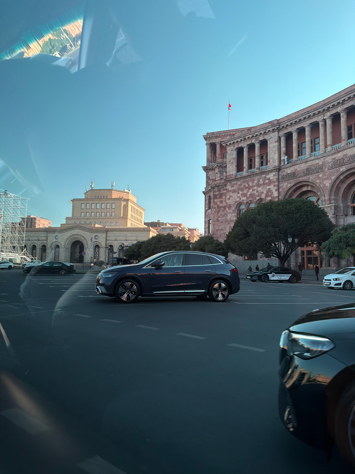 The image depicts a street scene in Yerevan, Armenia, likely Republic Square. In the foreground, a dark-colored SUV is driving on the road. To the right, a portion of another dark-colored car's front end is visible. In the mid-ground, there are several other vehicles, including a black sedan with markings that appear to be a police vehicle, and a white sedan. A large, ornate building with classical architecture, characterized by columns and arches, dominates the right side of the frame. This building is likely the National Gallery of Armenia or the Government House. To the left of this building is another, lighter-colored building with a distinctive facade and antennas on its roof, possibly the Ministry of Foreign Affairs. Trees line the area in front of the buildings. The sky is clear and blue, suggesting a daytime setting. A flag is visible on a flagpole atop the classical building. The scene is captured from a moving vehicle, as evidenced by reflections on the windshield.