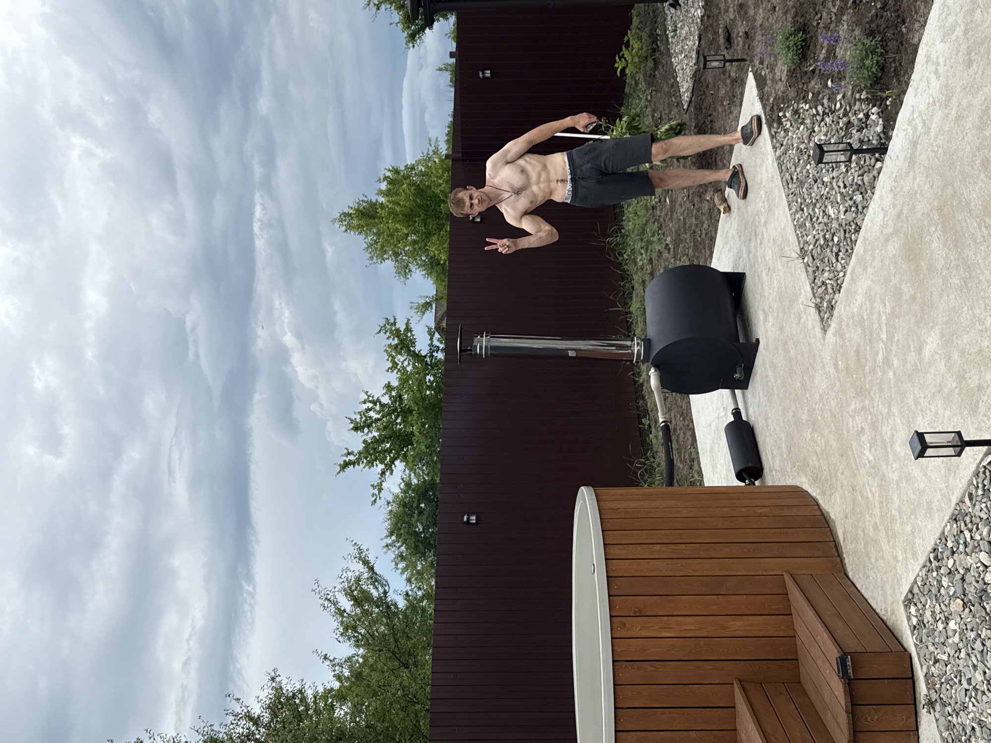 The image depicts an outdoor scene in Starokorsunskaya, Russia, featuring a wooden hot tub with integrated steps on the left. Adjacent to the hot tub is a black wood-fired stove with a stainless steel chimney. A shirtless man, exhibiting a lean physique and wearing dark shorts and sandals, stands near the stove on the right, making a peace sign with his left hand. He is holding a dark object in his right hand. The ground is a mix of concrete pathways and gravel landscaping. A dark brown wooden fence encloses the area, with lush green trees visible beyond it. The sky is overcast with numerous clouds. The scene appears to be a backyard setup for relaxation.