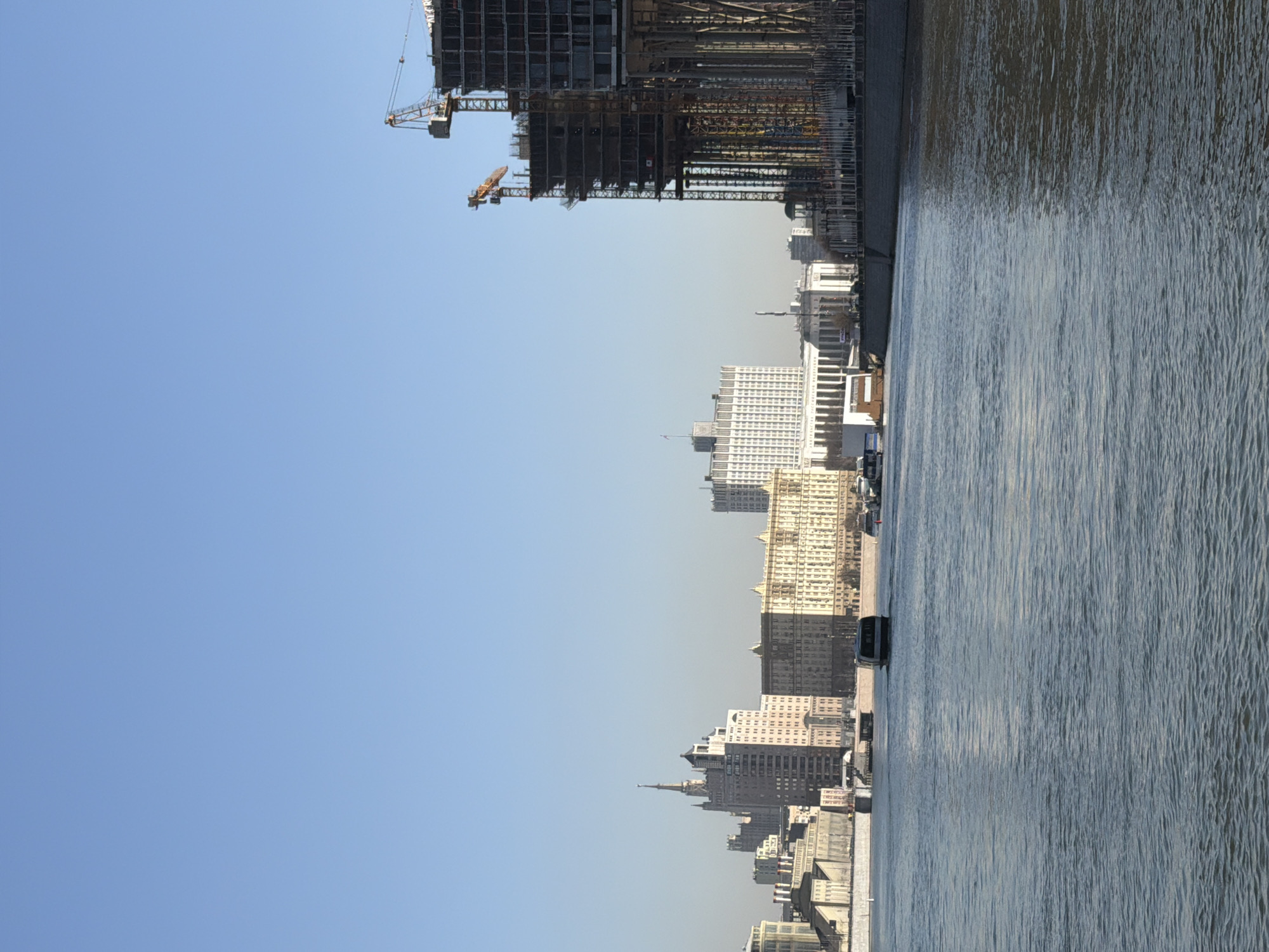 The image displays a daytime, panoramic view across the Moskva River in Moscow, Russia, towards the city's urban landscape. The lower half of the frame is occupied by the Moskva River, its surface showing light ripples. A dark, rectangular riverboat or ferry is observed mid-river, positioned towards the center-left, moving against the flow.
Along the far bank, a grey concrete embankment lines the river. Above the embankment, a series of buildings form the Moscow skyline. The prominent, multi-story white building with a Russian flag flying from its roof, centrally located in the distant skyline, is identifiable as the Government House of the Russian Federation (White House). To the left of this building, the distinctive spire of the Kudrinskaya Square Building is visible among other city structures.
To the right, a significant portion of the image is dominated by a large, multi-story building under active construction. This structure presents an exposed concrete framework and extensive scaffolding. Two large, orange and white tower cranes are positioned on and above the building, indicating ongoing development. The sky above is clear and light blue, suggesting fair weather conditions. No individuals are discernible in the scene.
