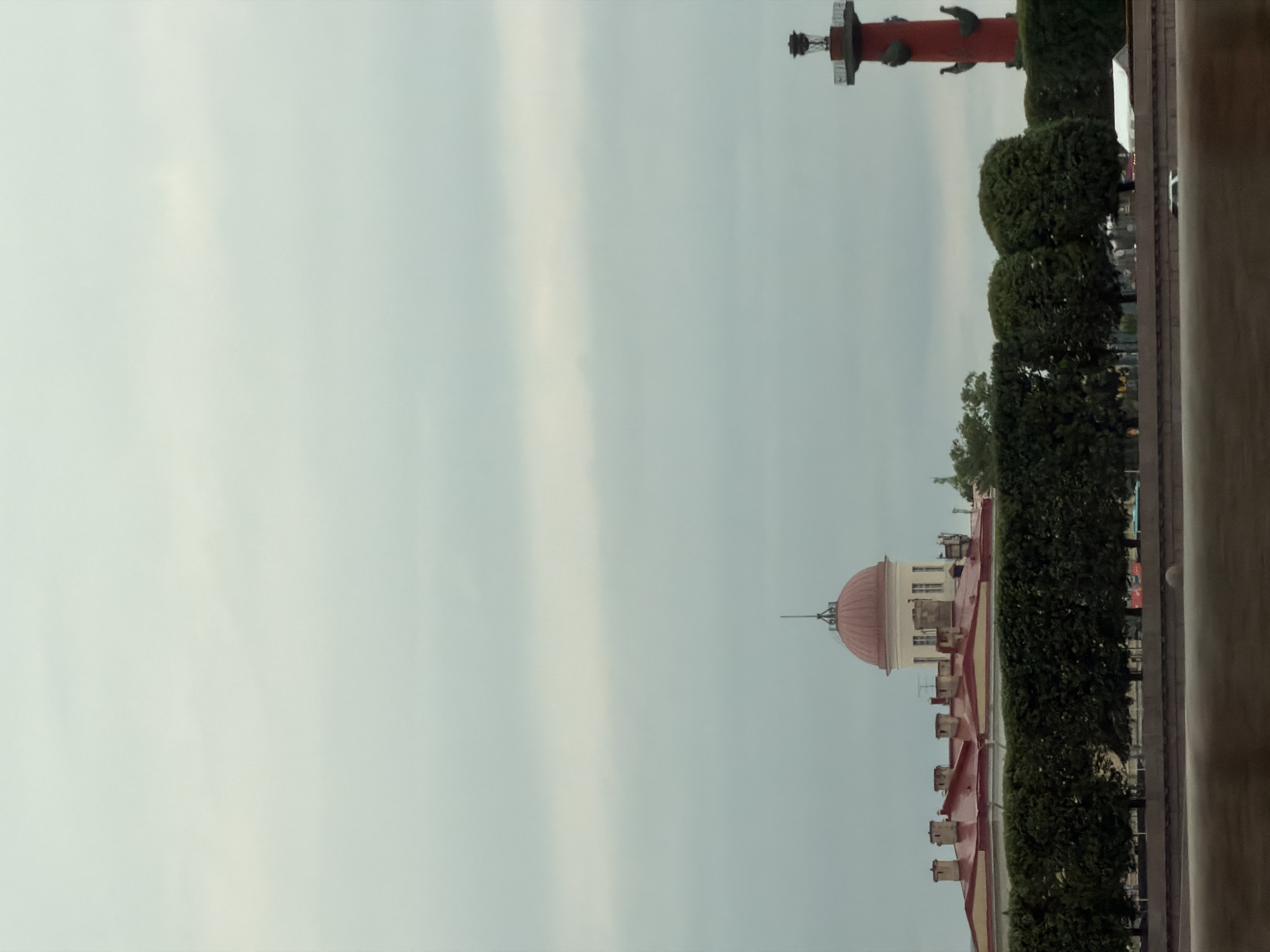 The image depicts an overcast sky above a cityscape. In the middle ground, a row of manicured green trees forms a lush barrier. Behind the trees, a long, red-roofed building with a dome topped by a spire is visible, flanked by chimneys. To the right of the building, a tall, red column, likely a monument or landmark, stands prominently. The foreground features a body of water, possibly a river or canal, with a blurred, reflective surface. The overall scene suggests a view of an urban architectural ensemble under a cloudy sky.