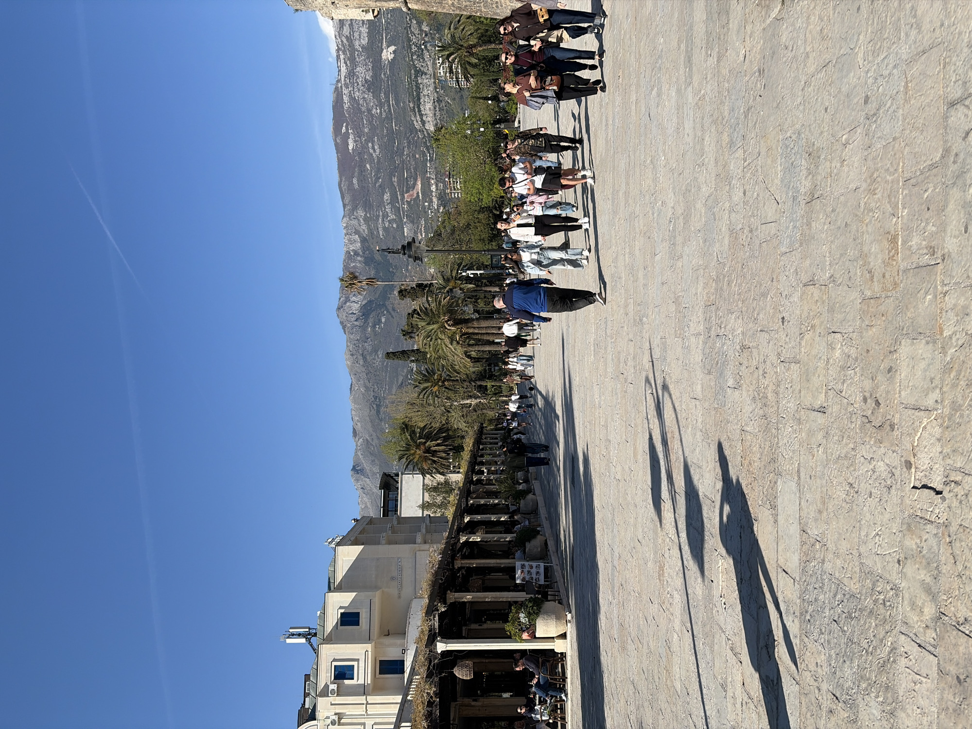 The image depicts a street scene in Budva, Montenegro. The sky is clear and blue. A group of people walks along a stone-paved path, parallel to a line of palm trees and a building. The building is a light color with blue window frames. A mountain range rises in the background. The shadows of people and lampposts stretch across the stone path.