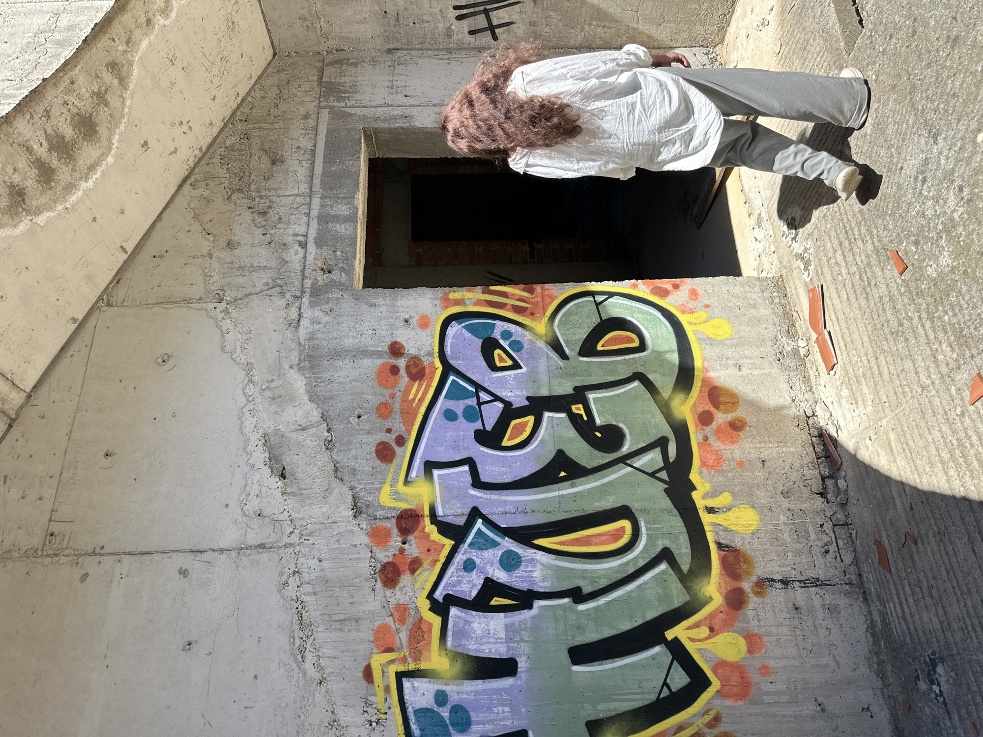 An interior view of an unfinished or abandoned concrete structure, perhaps in Gornji Katun, Montenegro. Graffiti adorns the lower portion of a wall, and a person, wearing a white shirt and gray pants, stands near an open, window-like space. The person is facing away from the viewer, their hair is brown-red.
