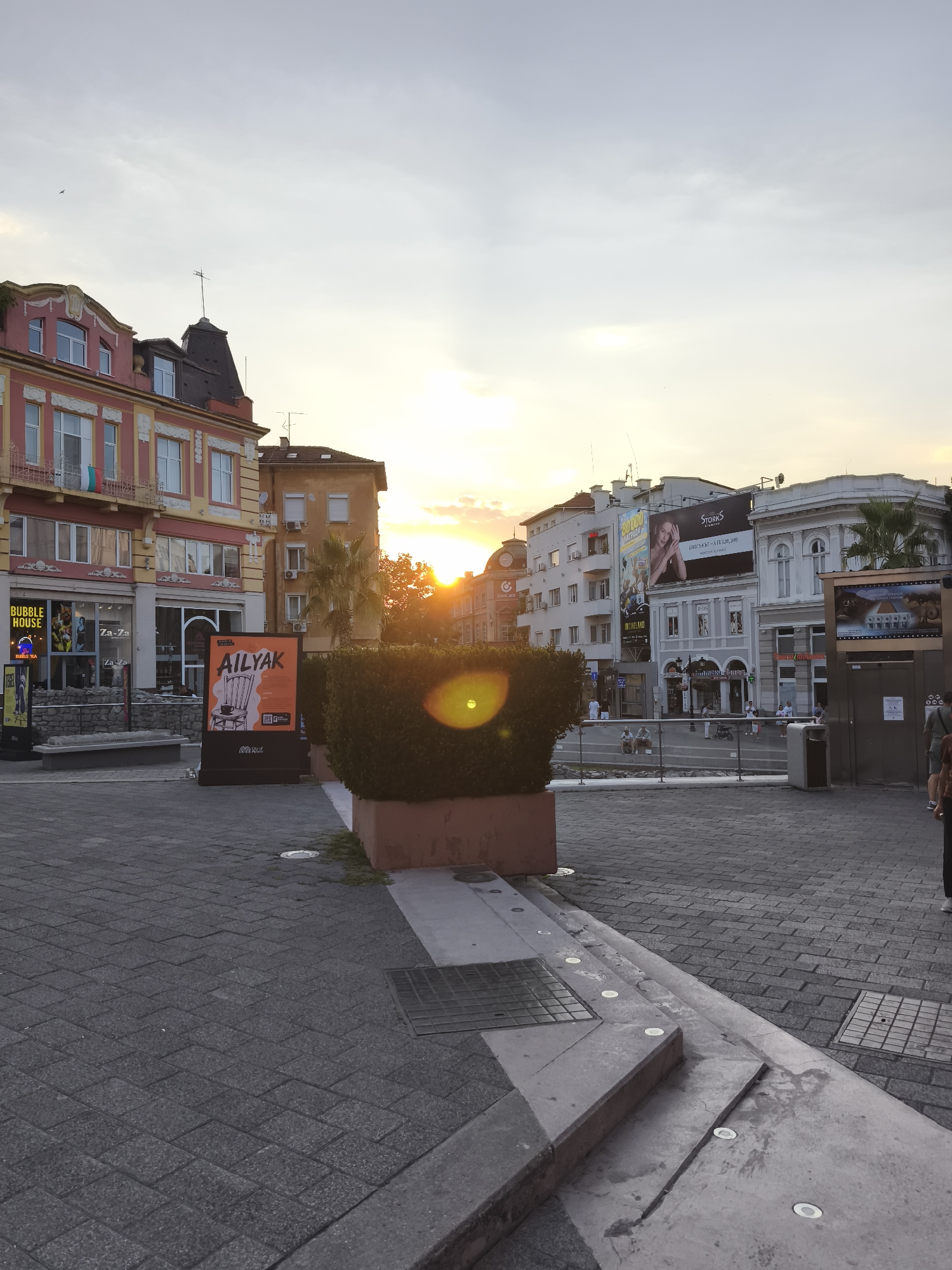 The image shows a street scene in the city of Plovdiv, Bulgaria, during sunset. The sun is low in the sky, casting a warm glow and creating lens flare in the foreground. Buildings line the street, featuring a mix of architectural styles, some with colorful facades and ornate details, while others are more modern with large billboards. People are visible walking and sitting on benches, going about their evening. A prominent hedge in the foreground partially obstructs the view of the street and buildings. Paved sidewalks and a cobblestone street are visible. The overall impression is of a vibrant urban environment at the close of day.