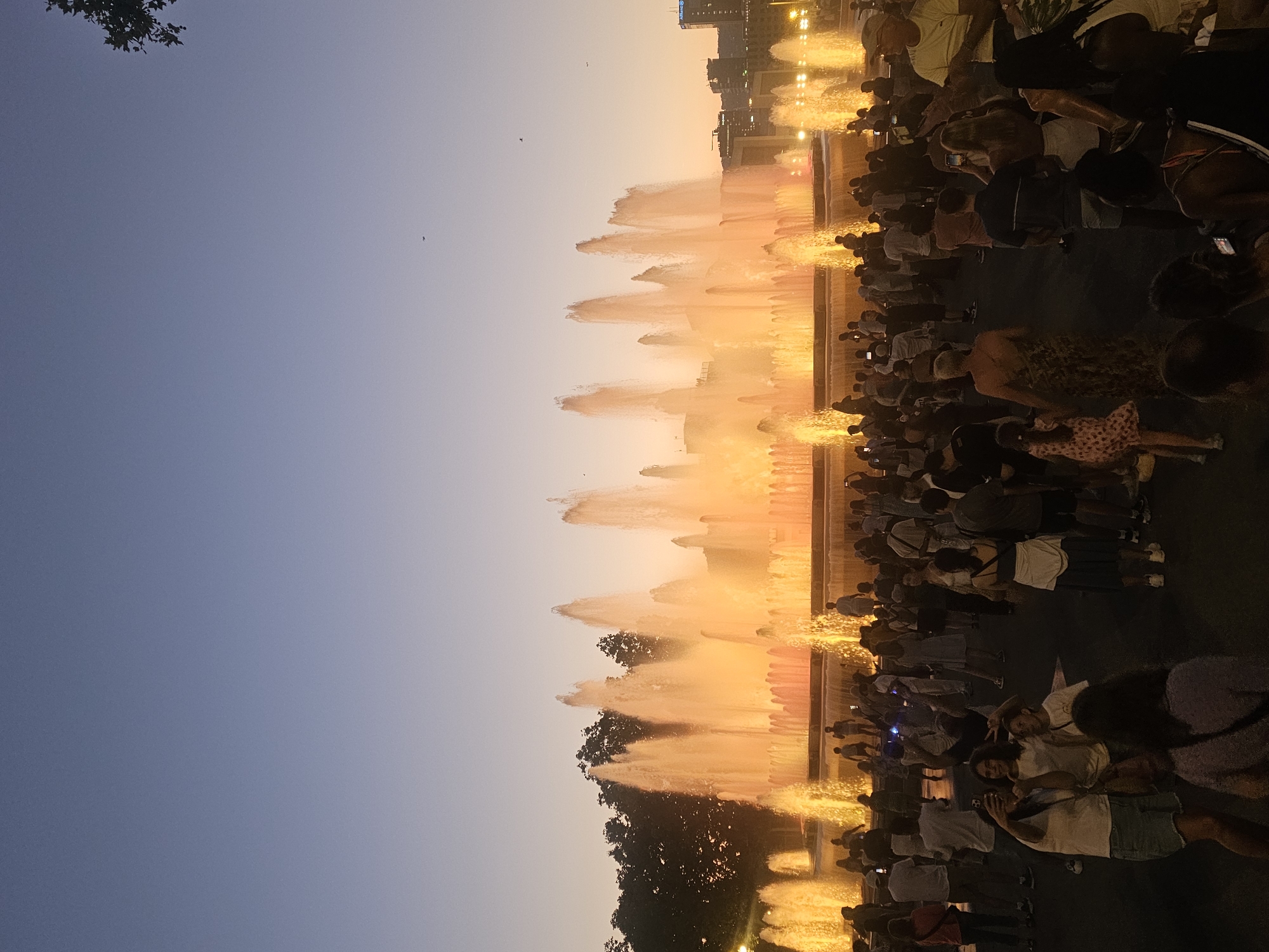 The image depicts a wide shot of the Magic Fountain of Montjuïc in Barcelona, Spain, at dusk. The fountain is illuminated with vibrant orange and yellow lights, creating dramatic water displays. A large crowd of people is gathered in front of the fountain, many of whom are taking pictures or videos with their phones. The sky above is a gradient of deep blue to a lighter hue near the horizon, with a few birds visible. Trees line the background behind the fountain, and some buildings can be seen on the right side of the image, including one with an "Allianz" sign. The atmosphere appears festive and lively, with people of all ages enjoying the spectacle.