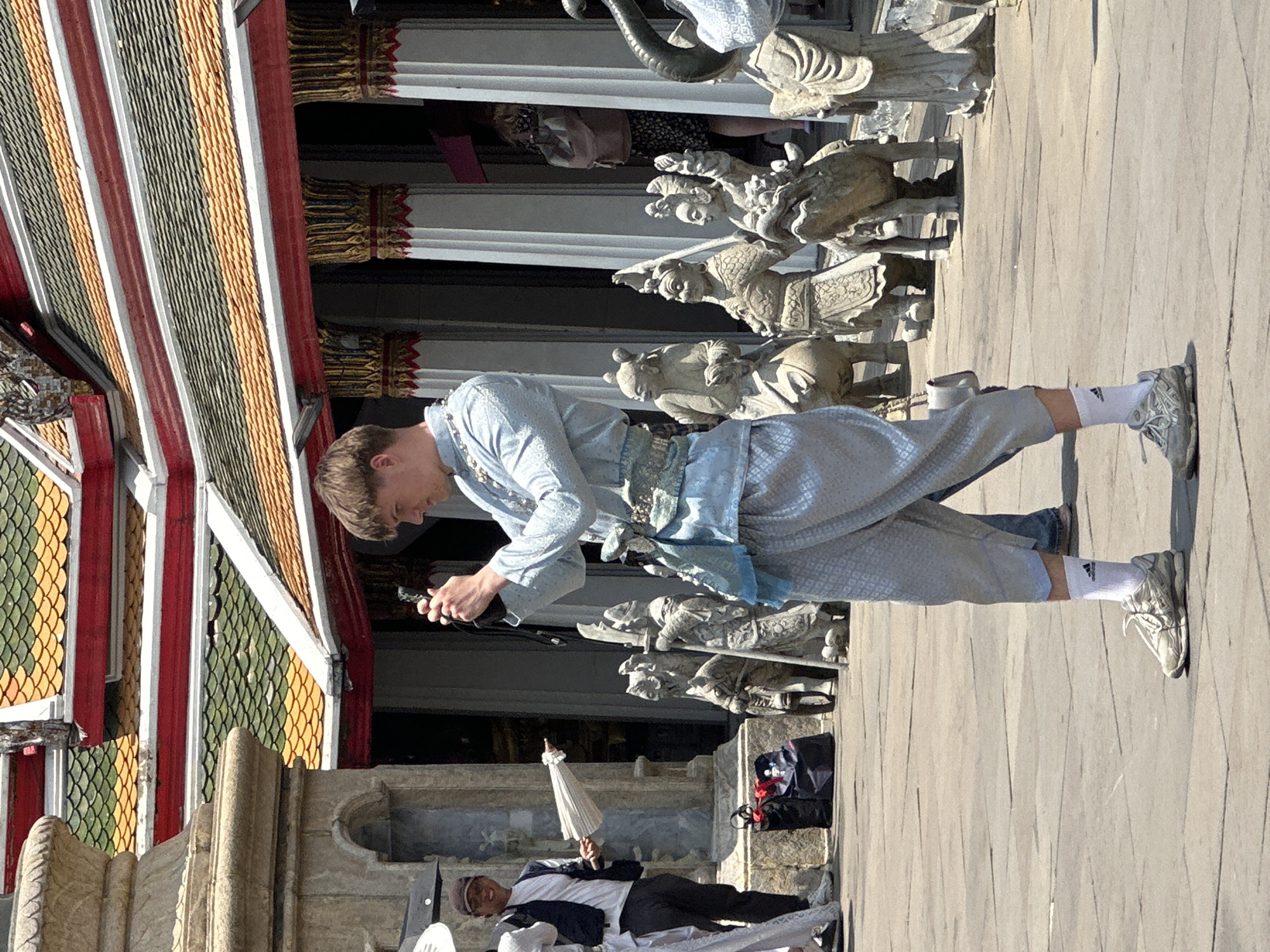 The image shows a man standing on a paved surface next to a temple in Bangkok, Thailand. He is dressed in a traditional light blue Thai outfit and is holding a dark object in his hands, possibly a phone or camera. The temple features a multi-tiered roof with decorative gold elements and red and white trim. A row of stone statues lines the edge of the temple. Another person lies on the ground nearby, and another person can be seen behind the statue. The environment appears to be sunny and outdoors, with the temple structure providing a backdrop.