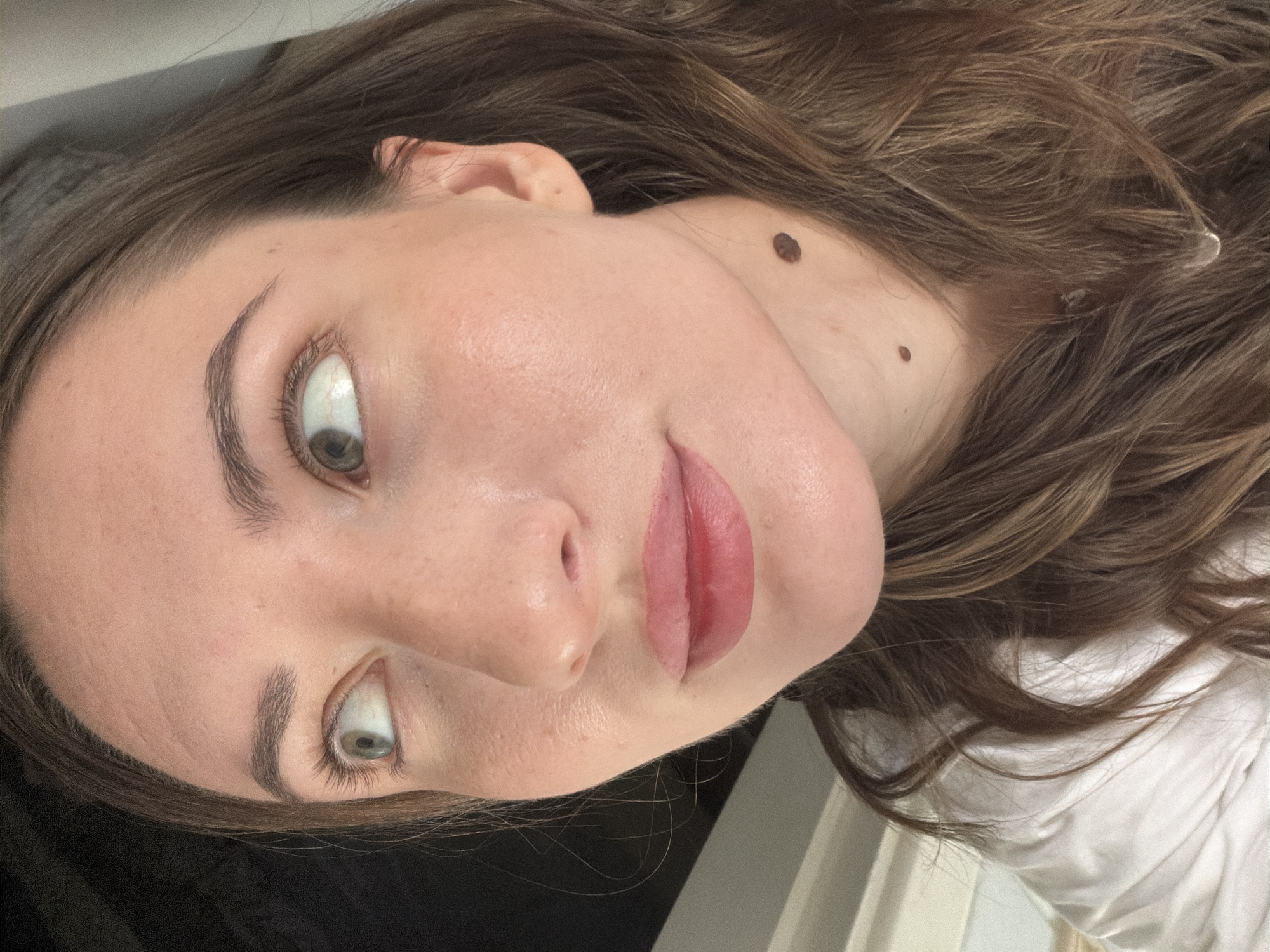 The image is a close-up, waist-up shot of a young woman's face and upper body. She has light skin, brown hair, and is looking to her left. She is wearing a white shirt. Two moles are visible on her neck, one slightly higher than the other. The background is dark with some vertical lines suggesting a window frame or blinds. The lighting appears natural and originates from the right. The image does not depict any specific location in Sharm El Sheikh, Egypt.
