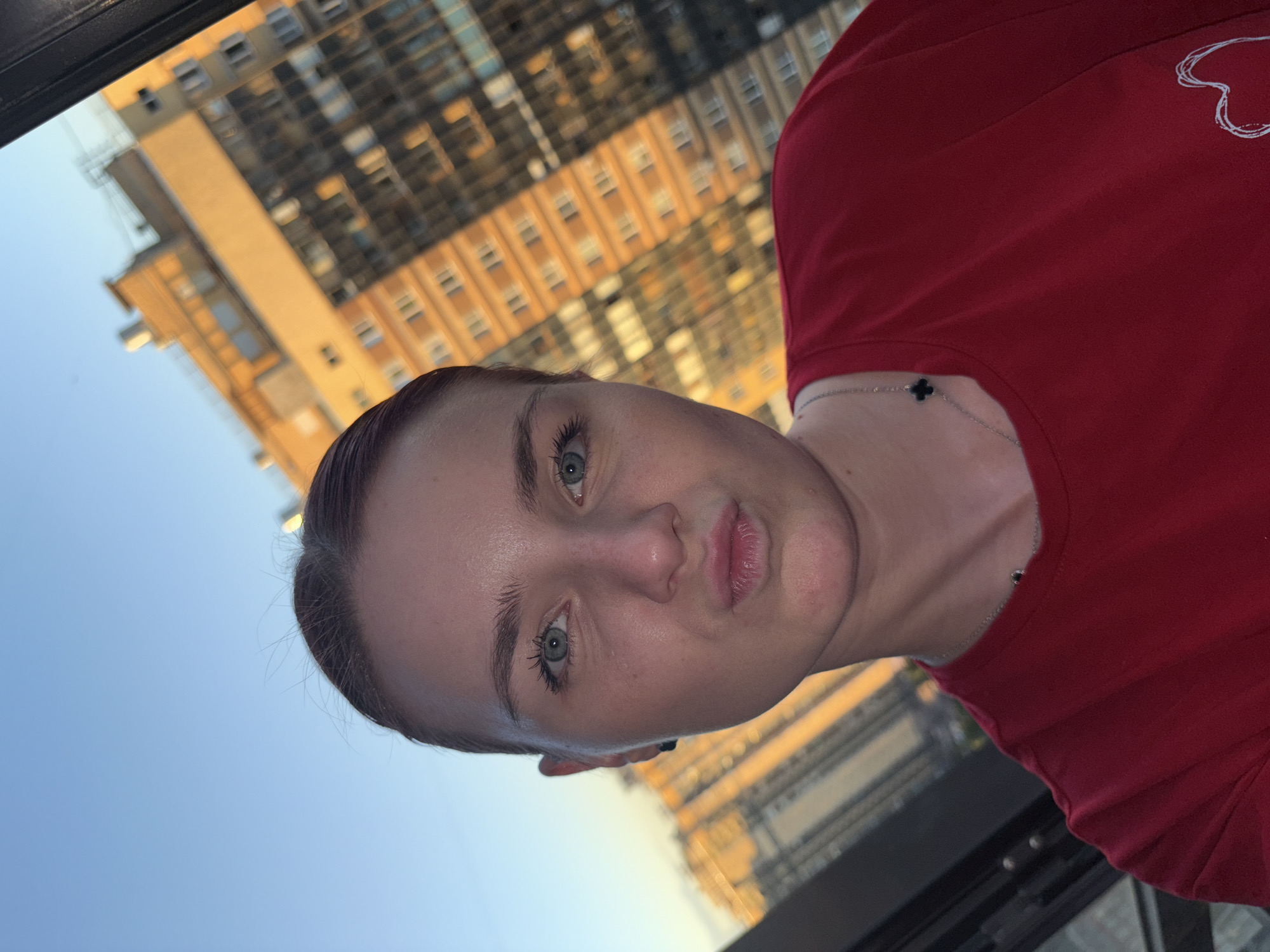 The image is an outdoor, low-angle, waist-up shot of a young woman with light skin and light blue eyes. She is wearing a red t-shirt, and a delicate silver necklace with a dark clover-shaped charm. Her hair is dark brown and pulled back, revealing her forehead and ears. Her expression is a slight pout, with her lips pursed.
Behind her, a large, modern building with many windows is visible, appearing to be illuminated by the setting sun, suggesting it is late afternoon or early evening. The sky above is a clear blue, indicating fair weather. The overall impression is casual, possibly a selfie taken on a balcony or from a high vantage point, with the urban landscape serving as a backdrop.