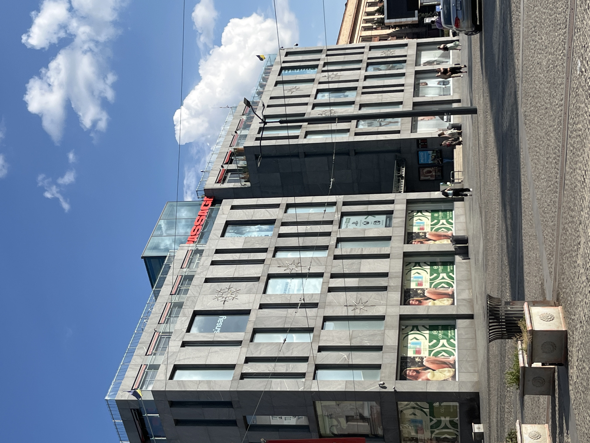 The image shows a modern, multi-story building with a gray stone facade. The building has many windows and some signage. On one section of the building, red lettering spells out "SINSY". Another sign features the word "MOTO". There are also advertisements displayed in some of the windows. One advertisement shows people with the text "COLLIBANK" above it.
Several people are visible in the image. Some are walking on the sidewalk in front of the building, while others appear to be sitting or standing near the entrance. One person is seen walking a dog. Another person is standing near a car parked on the street.
The scene appears to be a public space, possibly a commercial district or a city center. The ground is paved with cobblestones, and there are tram tracks visible. The sky is blue with some white clouds. The lighting suggests it is daytime, likely late morning or afternoon. A small flag, yellow and blue, is flying from a pole on the building. The overall atmosphere seems to be one of urban activity and daily life. The time of day is illuminated by bright sunlight, casting distinct shadows.