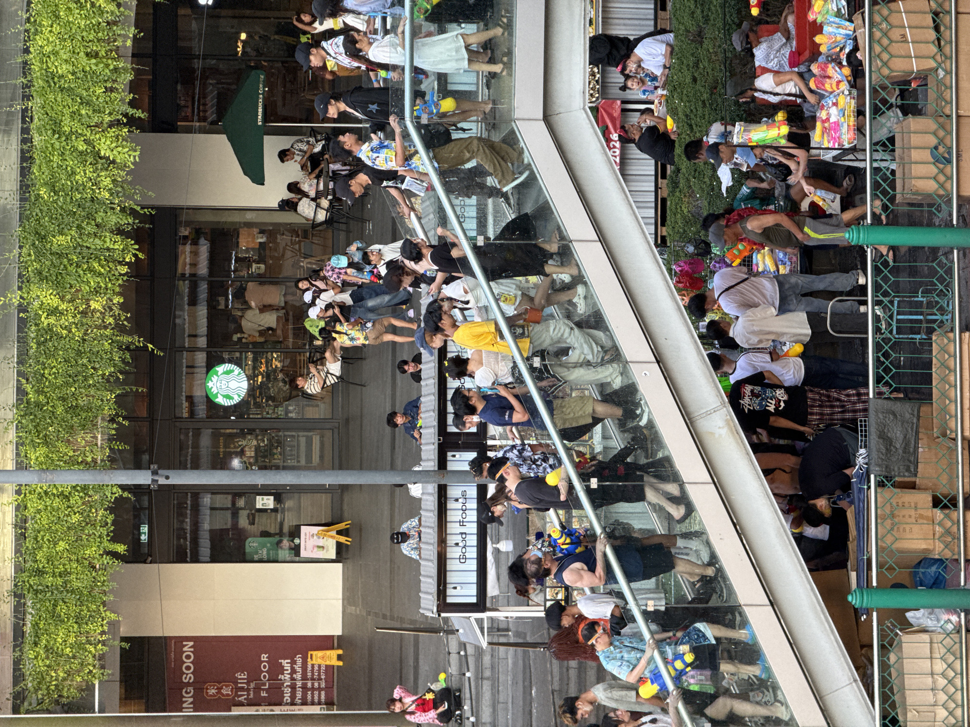 The image depicts a street scene in Bangkok, Thailand, likely during a Songkran water festival. A large crowd of people, many armed with water guns, are visible on both sides of an elevated walkway. The crowd includes people of diverse ages and appearances, some wearing festive clothing or carrying water containers. Some are engaged in playfully spraying water at each other. The elevated walkway is constructed with glass panels and metal supports.
In the background, the exterior of a building is visible, with a Starbucks coffee shop identifiable by its logo and green awning. Signage on the building also indicates a "COMING SOON" section and a "FLOOR" indication.
On the right side of the image, a green metal fence separates the crowd from a stack of cardboard boxes and various items for sale, possibly related to the festival. The overall atmosphere is lively and festive, indicative of the Songkran celebration.